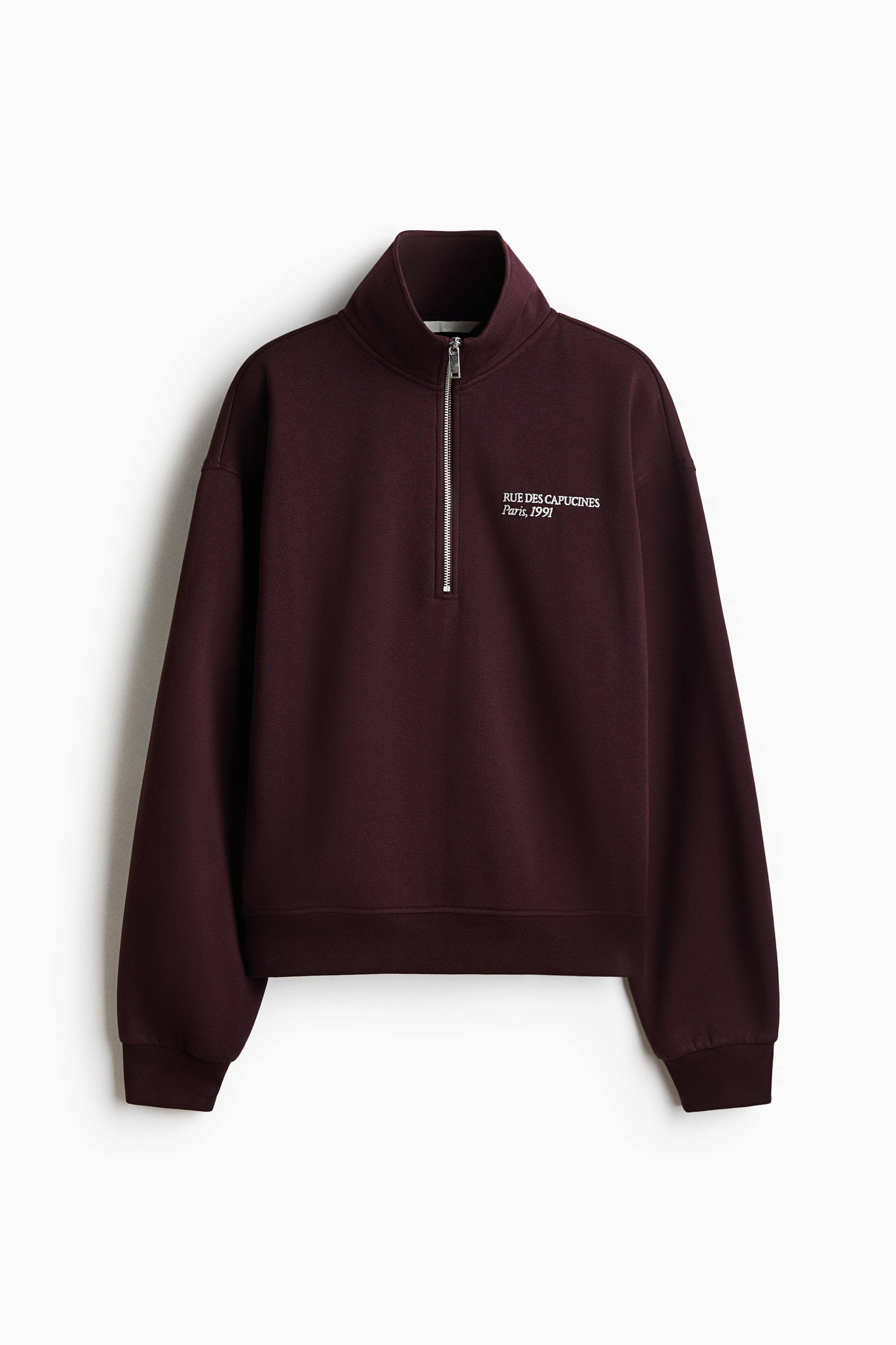 View larger image: A dark burgundy fleece sweatshirt is facing front, featuring a stand collar with a silver-tone quarter-zip, dropped shoulders, and ribbed cuffs and hem. The sweatshirt is displaying small, white printed text on the left chest that reads "RUE DES CAPUCINES Paris, 1991".