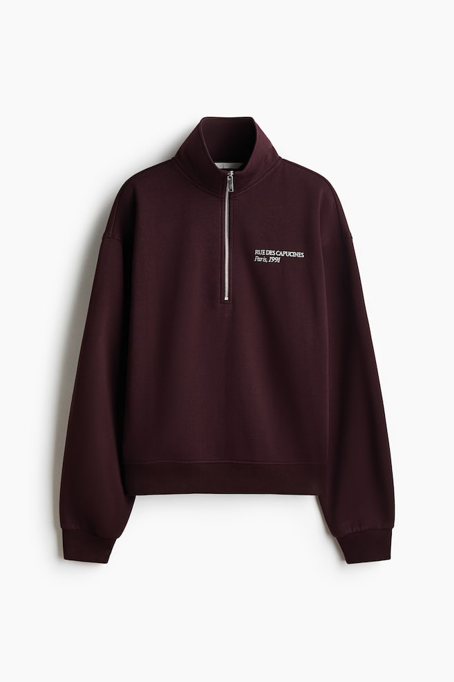 Ein dunkelburgunderfarbener Fleece-Sweatshirt ist von vorne zu sehen und verfÃ¼gt Ã¼ber einen Stehkragen mit silberfarbenem Quarter-Zip, Ã¼berschnittene Schultern sowie gerippte BÃ¼ndchen und einen gerippten Saum. Auf der linken Brust ist ein kleiner, weiÃer Aufdruck zu sehen, der âRUE DES CAPUCINES Paris, 1991â lautet. - 1