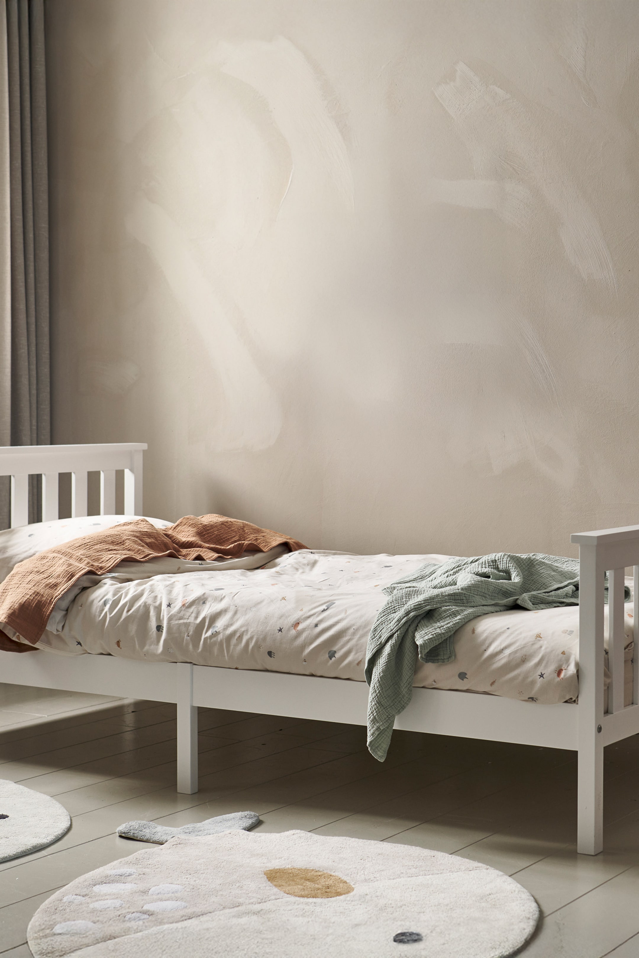 Agrandir l'image: Un cadre de lit en bois blanc présente une tête de lit et un pied de lit à lattes, positionnés à un léger angle vers la droite. Une couette de couleur claire avec de petits motifs colorés recouvre le matelas, avec une couverture texturée couleur rouille pliée à la tête et une couverture texturée vert clair drapée au pied.