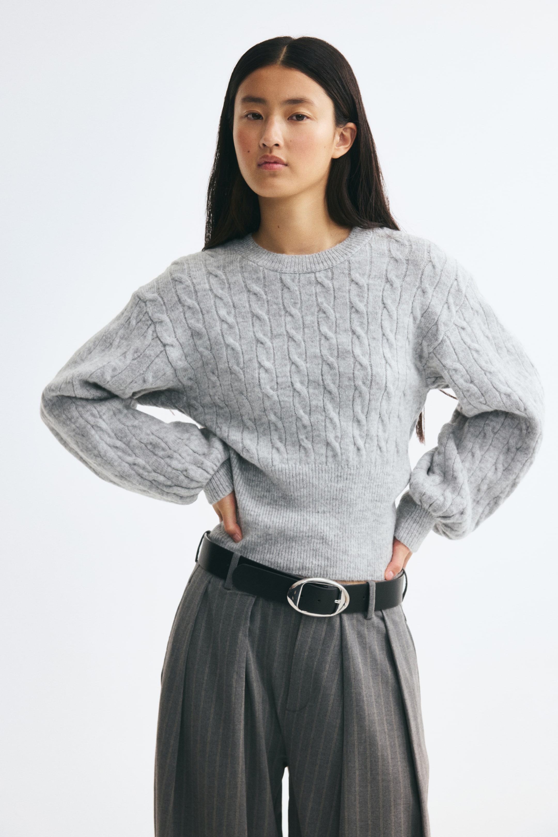 Größeres Bild anzeigen: Ein hellgrauer Zopfpullover wird von vorne gezeigt und verfügt über einen runden, gerippten Ausschnitt, überschnittene Schultern und lange Ärmel mit gerippten Bündchen. Der Pullover setzt einen breiten, gerippten Saum in Szene und ist hier mit grauen Nadelstreifen-Hosen und einem schwarzen Gürtel kombiniert.