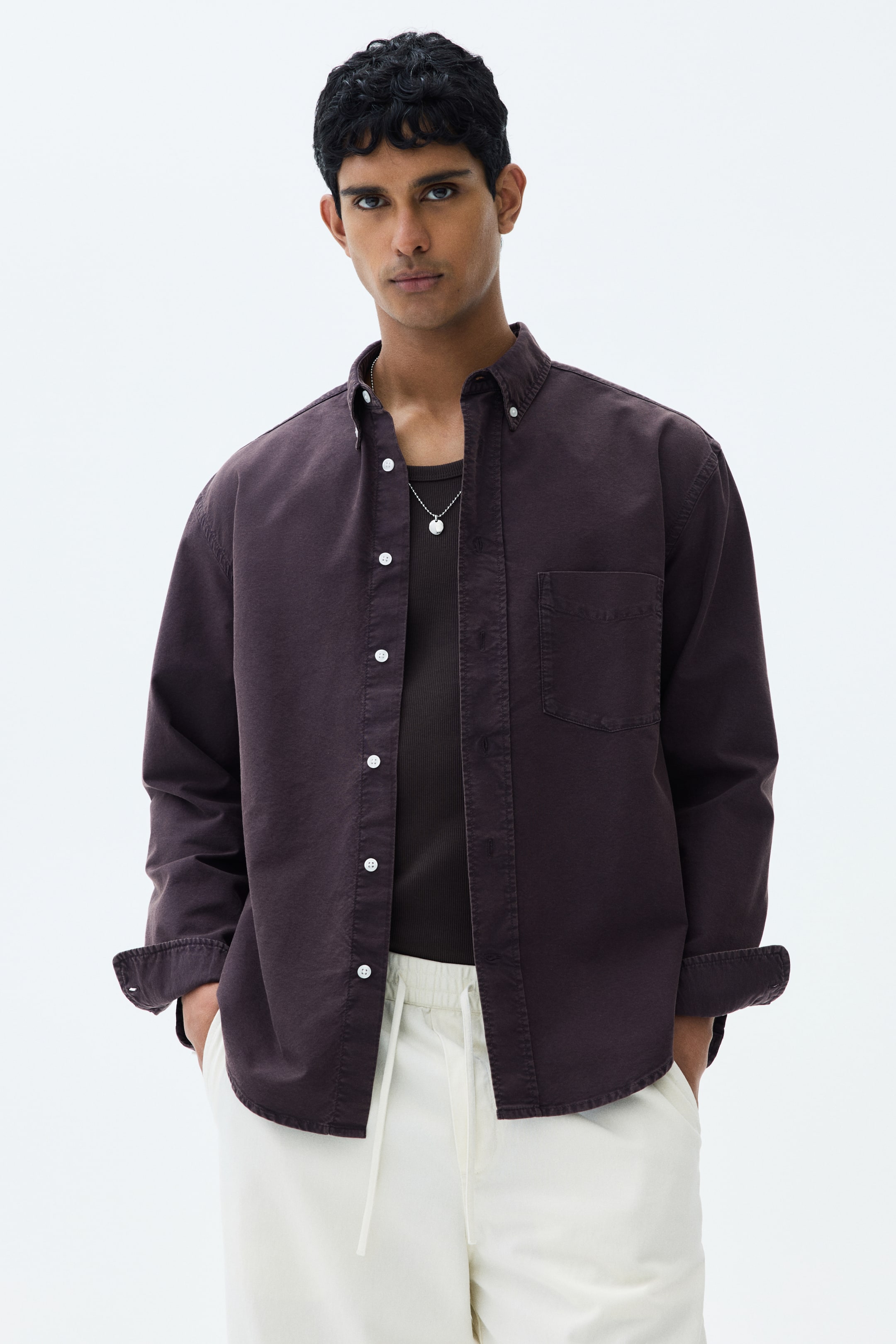 View larger image: The model is sporting a dark brown relaxed-fit shirt, featuring a button-down collar and an open chest pocket. The shirt is worn unbuttoned over a dark undershirt. Its long sleeves are casually rolled up, and the model's hands are comfortably placed in their pant pockets.