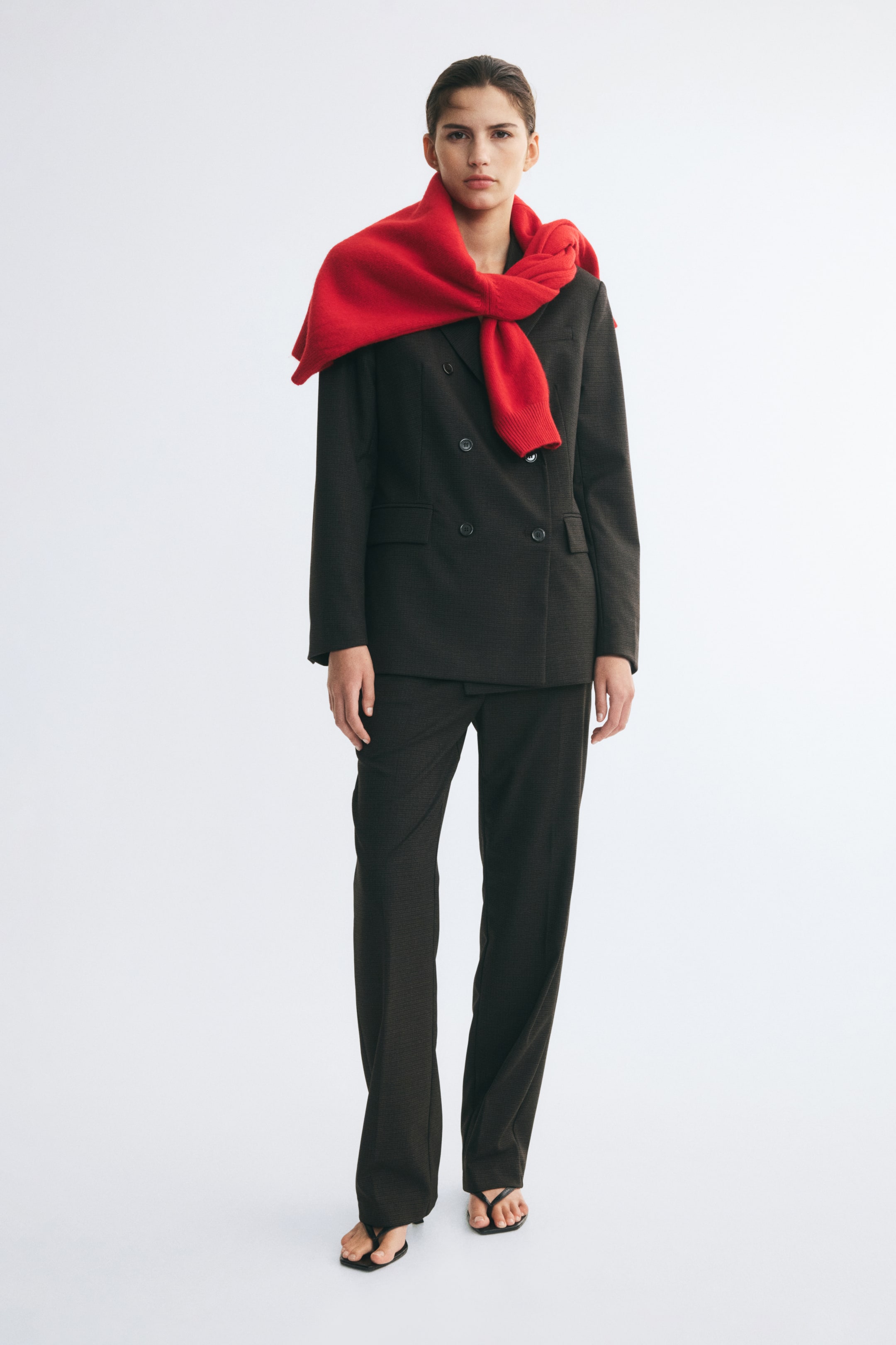 View larger image: Trousers in a dark woven fabric featuring straight legs with visible pressed creases are worn by the model. The trousers are styled as part of a suit with a matching double-breasted blazer and a bright red sweater draped over the shoulders.