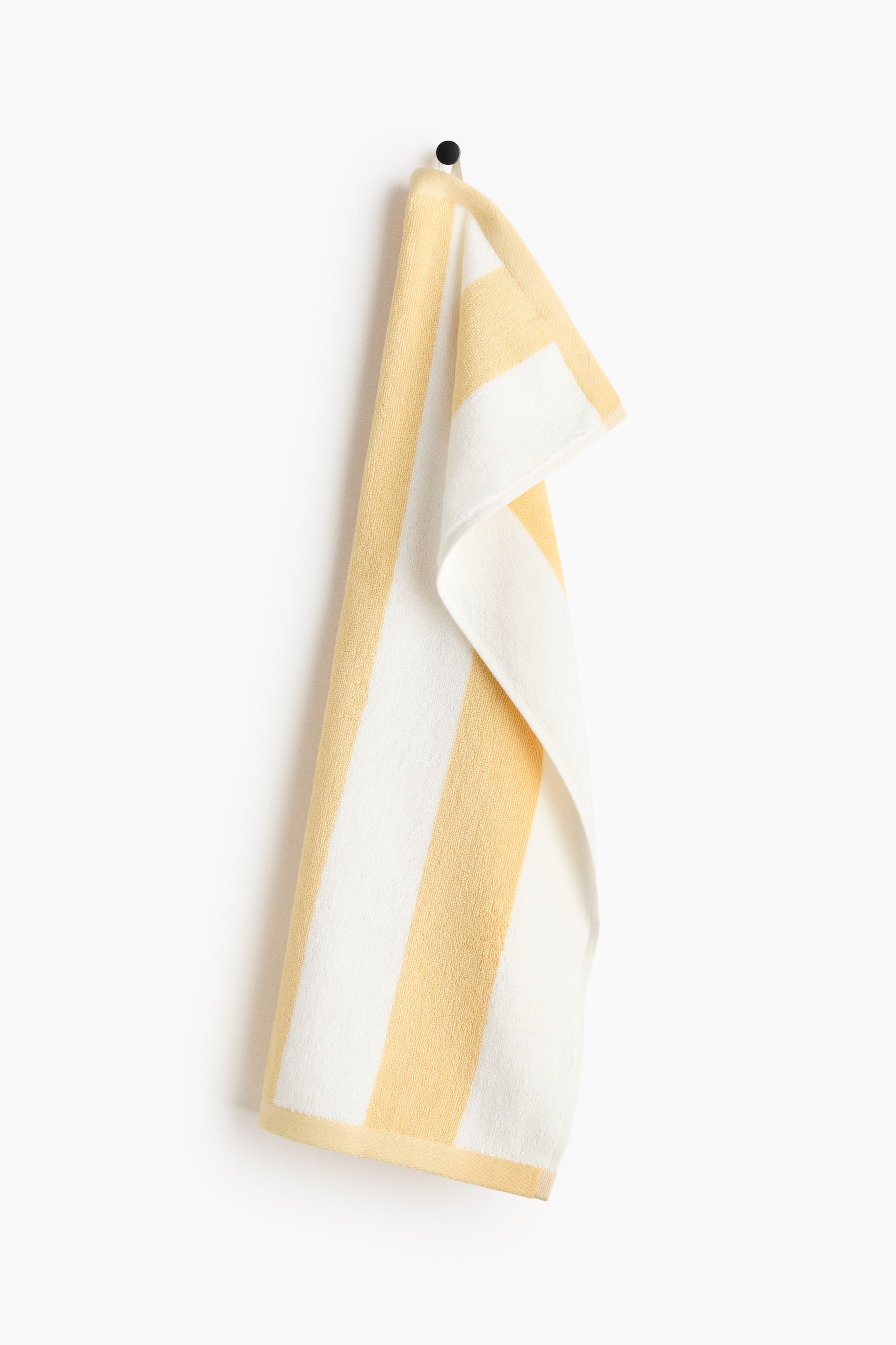 Büyük resmi görüntüle: Bir el havlusu, siyah bir kancadan asılı olarak sarı ve beyaz renklerin dönüşümlü dikey çizgilerini sergiliyor. Alt kenarında düz sarı bir dokuma kenar ile öne çıkıyor ve havlu kumaştan yapılmış.