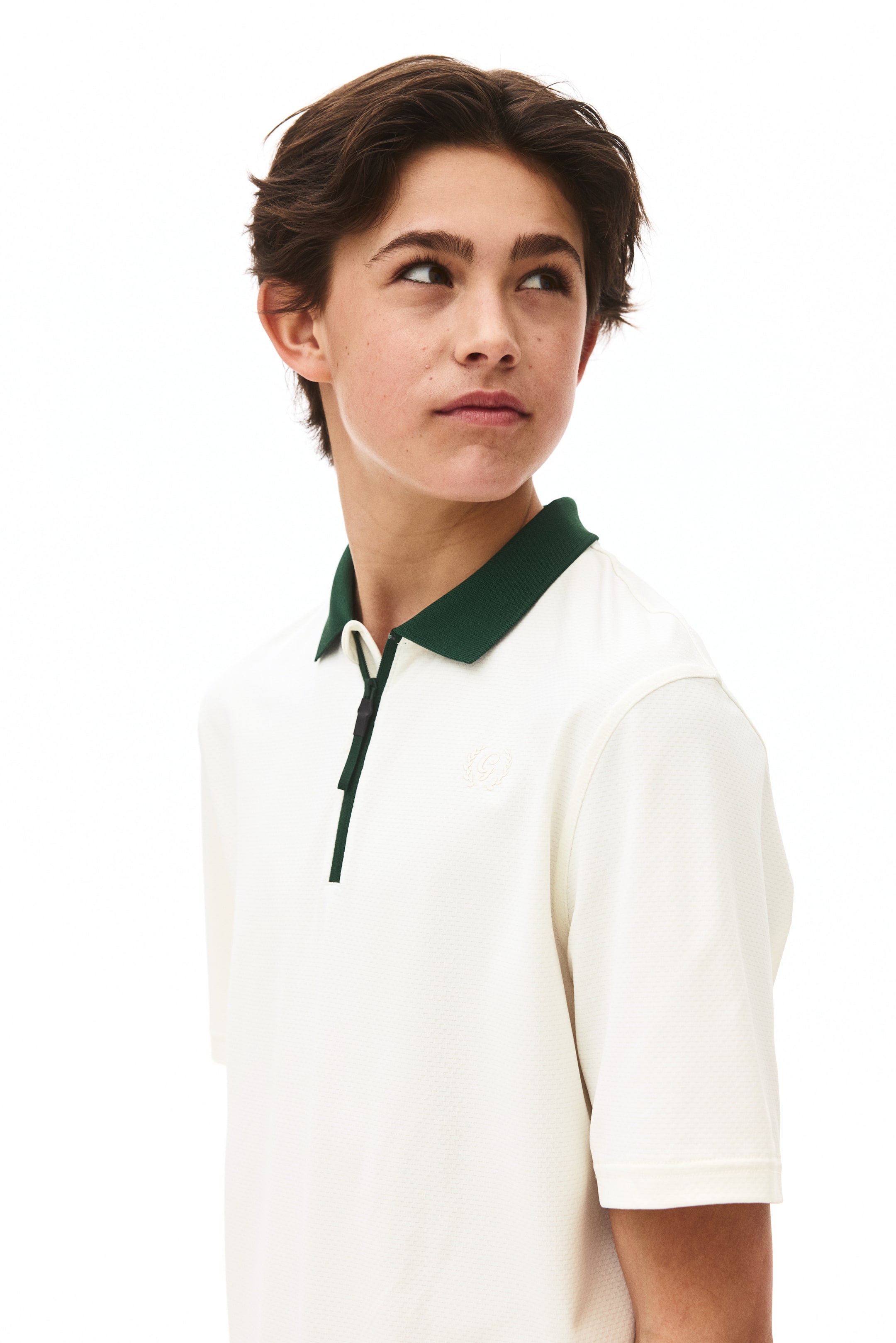 Visa större bild: En benvit pikétröja med korta ärmar bärs, med en mörkgrön krage, en kort mörkgrön knappslå och en diskret broderad logotyp i form av en vapensköld som syns på vänster bröst på dess texturerade tyg.