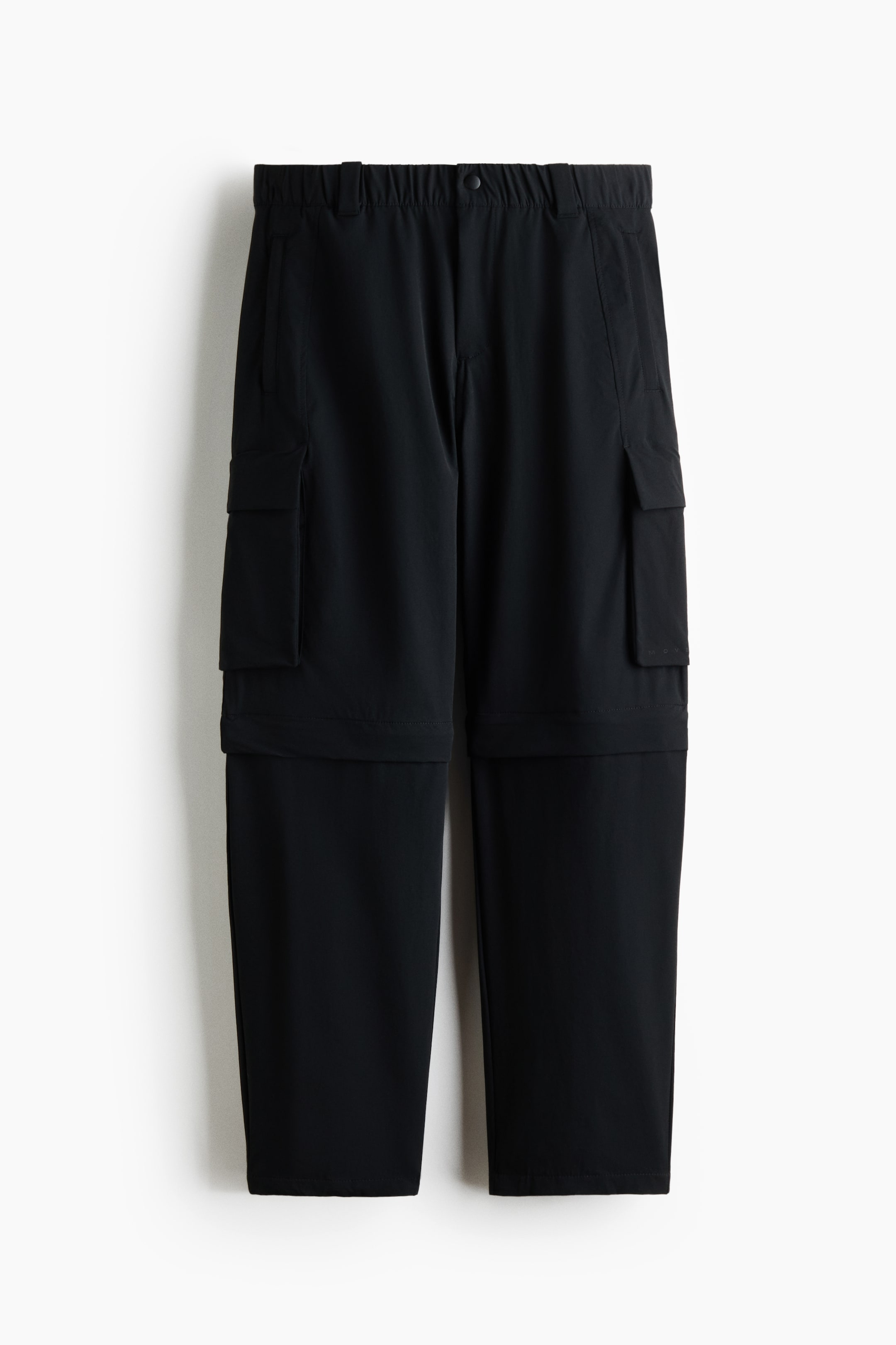Agrandir l'image: Pantalon convertible noir présenté de face, avec une taille élastiquée dotée de passants pour ceinture, une fermeture à bouton et deux poches fendues à l'avant. Deux poches cargo rectangulaires avec rabats sont visibles sur les cuisses extérieures, et les sections inférieures des jambes se rejoignent à la partie supérieure grâce à des fermetures éclair, formant ainsi un design convertible.