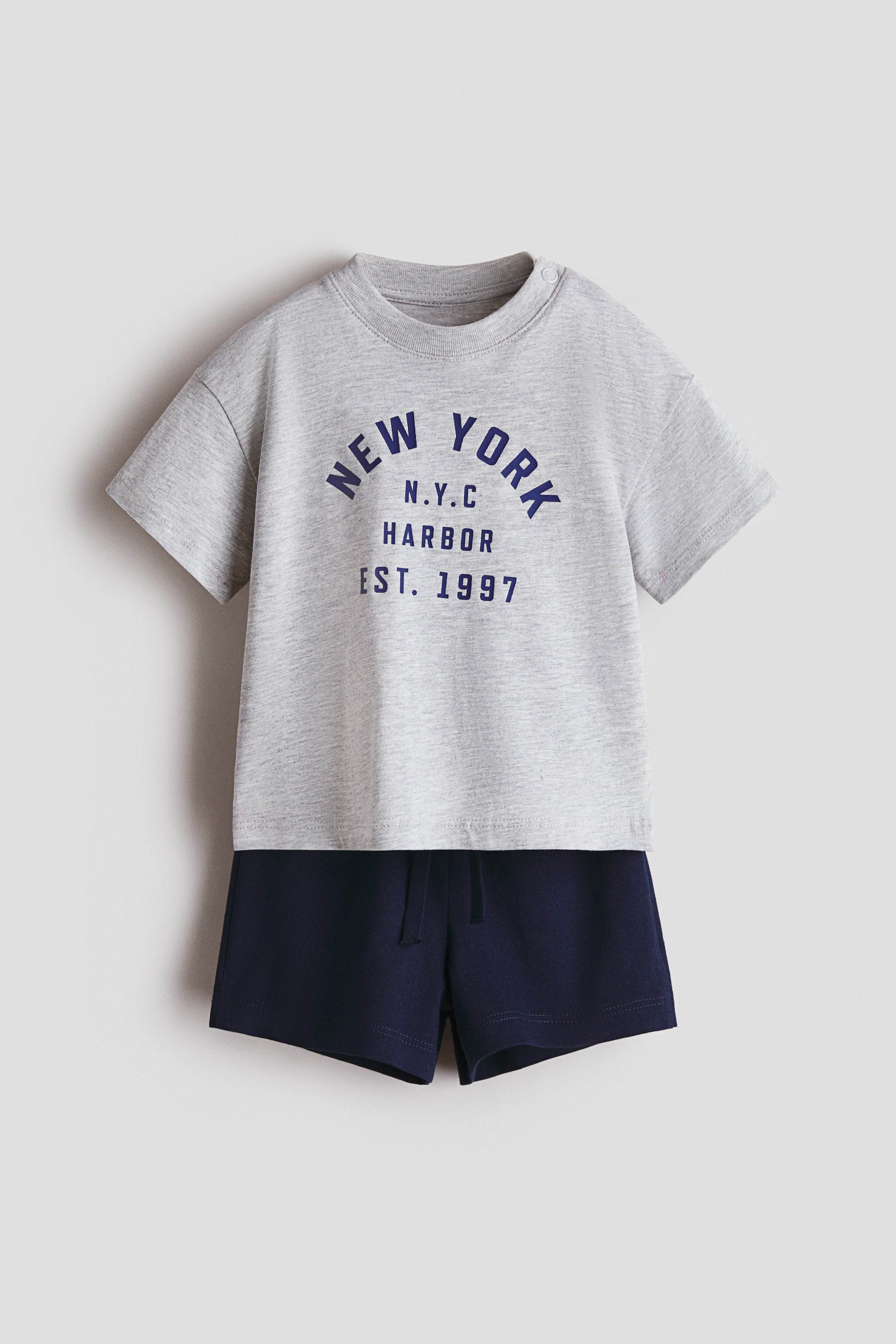 View larger image: A 2-piece cotton set includes a gray marl t-shirt and navy shorts, both shown from the front. The t-shirt features a ribbed crew neck, a visible snap button on one shoulder, short sleeves, and a dark blue "NEW YORK N.Y.C HARBOR EST. 1997" graphic on the chest. The shorts are designed with an elasticated drawstring waist.