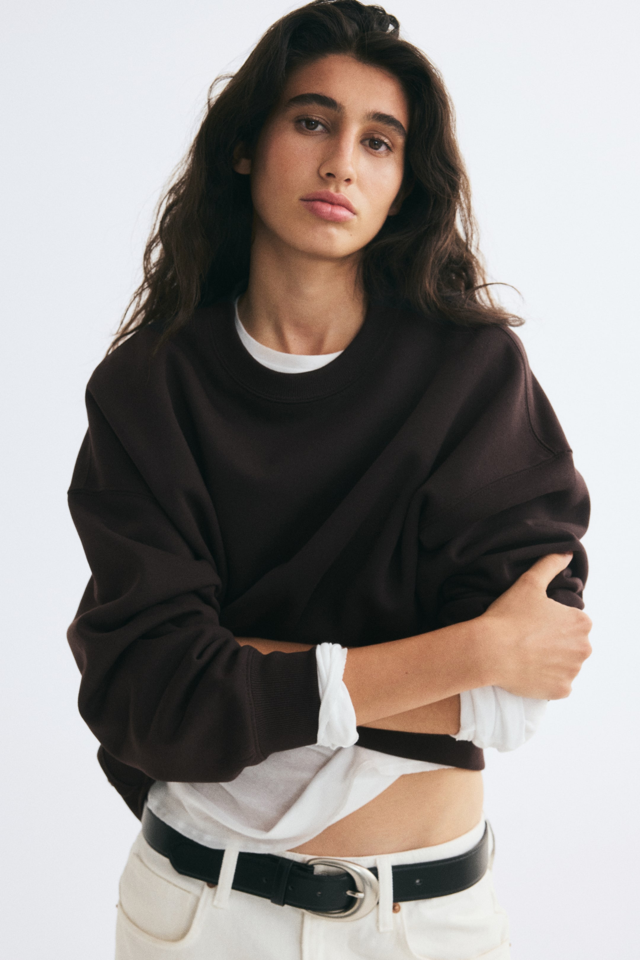 View larger image: An oversized dark brown sweatshirt with a crew neckline and dropped shoulders is worn by the model, who has their arms crossed. The sweatshirt is layered over a white T-shirt, which is visible at the neckline, cuffs, and hem.
