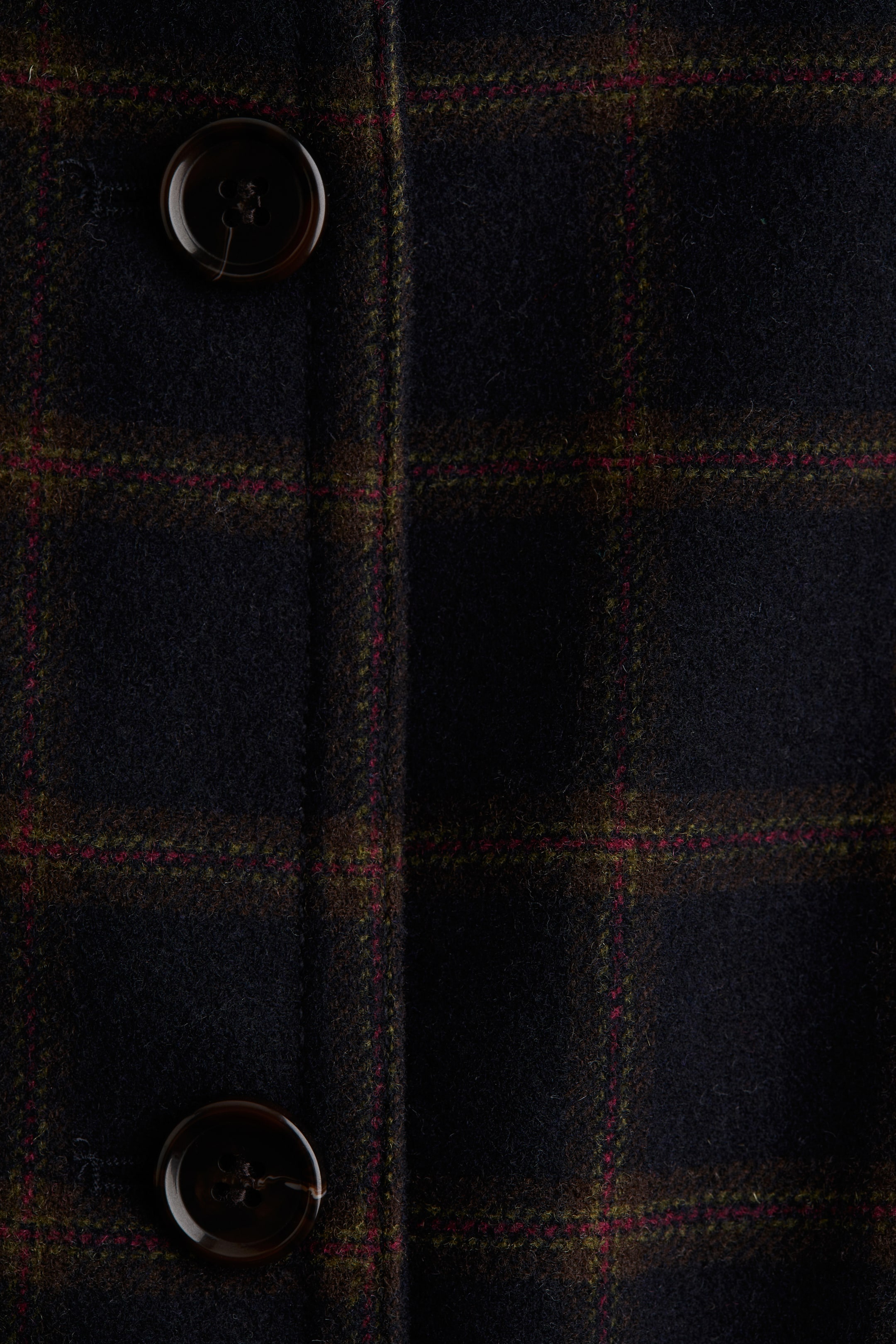 View larger image: A dark plaid woven fabric is featuring a pattern of olive green and red lines. Two dark brown round buttons are visible on a vertical placket, indicating a front closure.