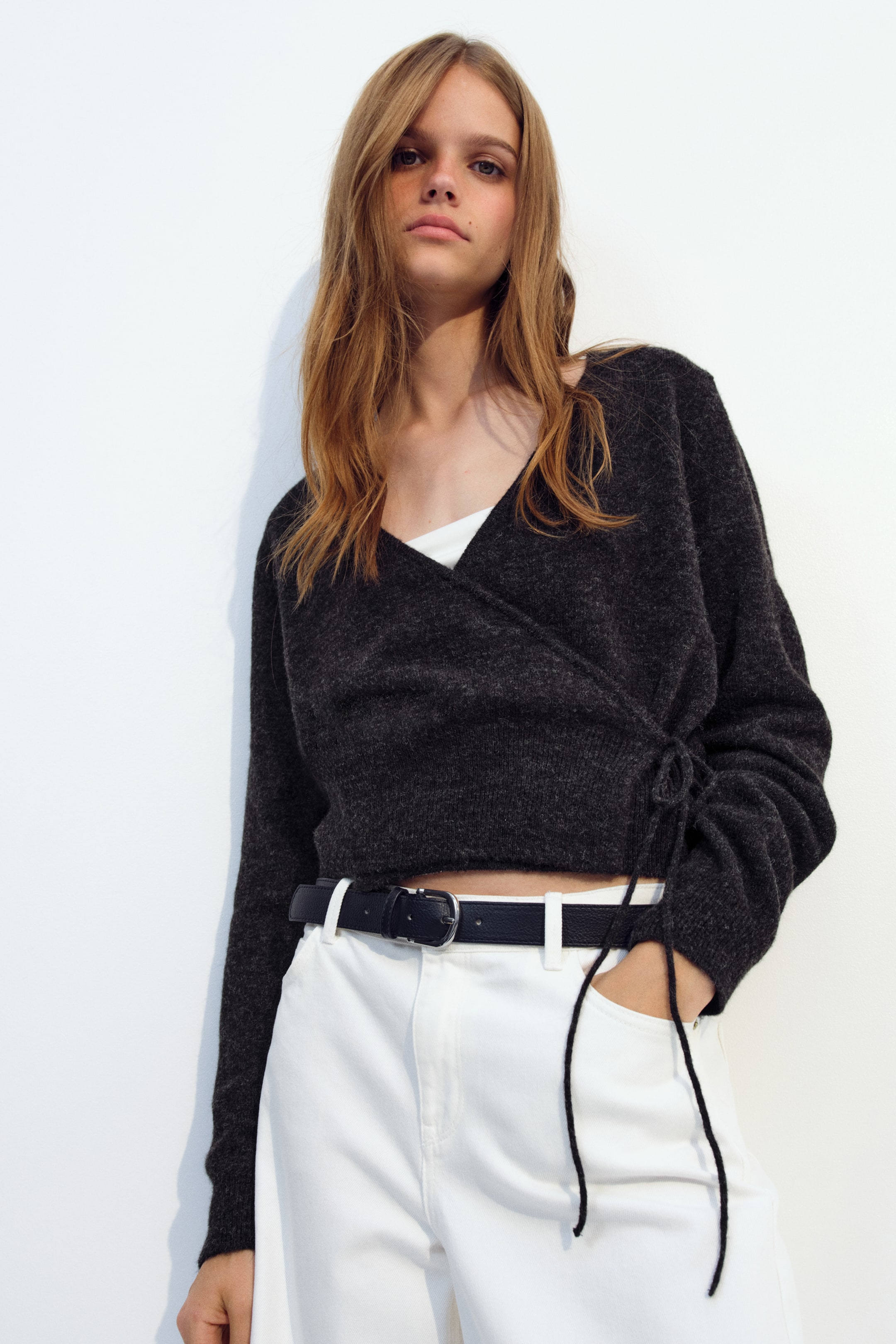 View larger image: The model is wearing a dark grey wrap cardigan in a soft knit, featuring a V-neckline, long sleeves, and a narrow side tie closure. The cardigan is layered over a white top and styled with white trousers and a black belt.