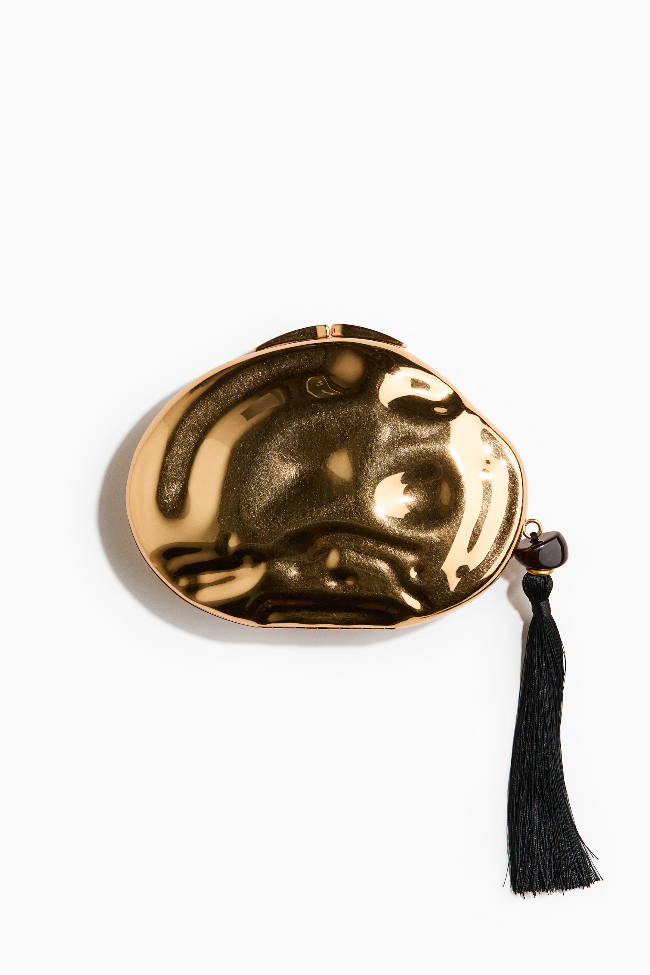 View larger image: "An asymmetric, golden metal clutch is showcasing a hammered finish with a reflective surface. At the top, a small purse clasp is visible, while on the right side, a black tassel, attached by a dark bead, elegantly hangs."