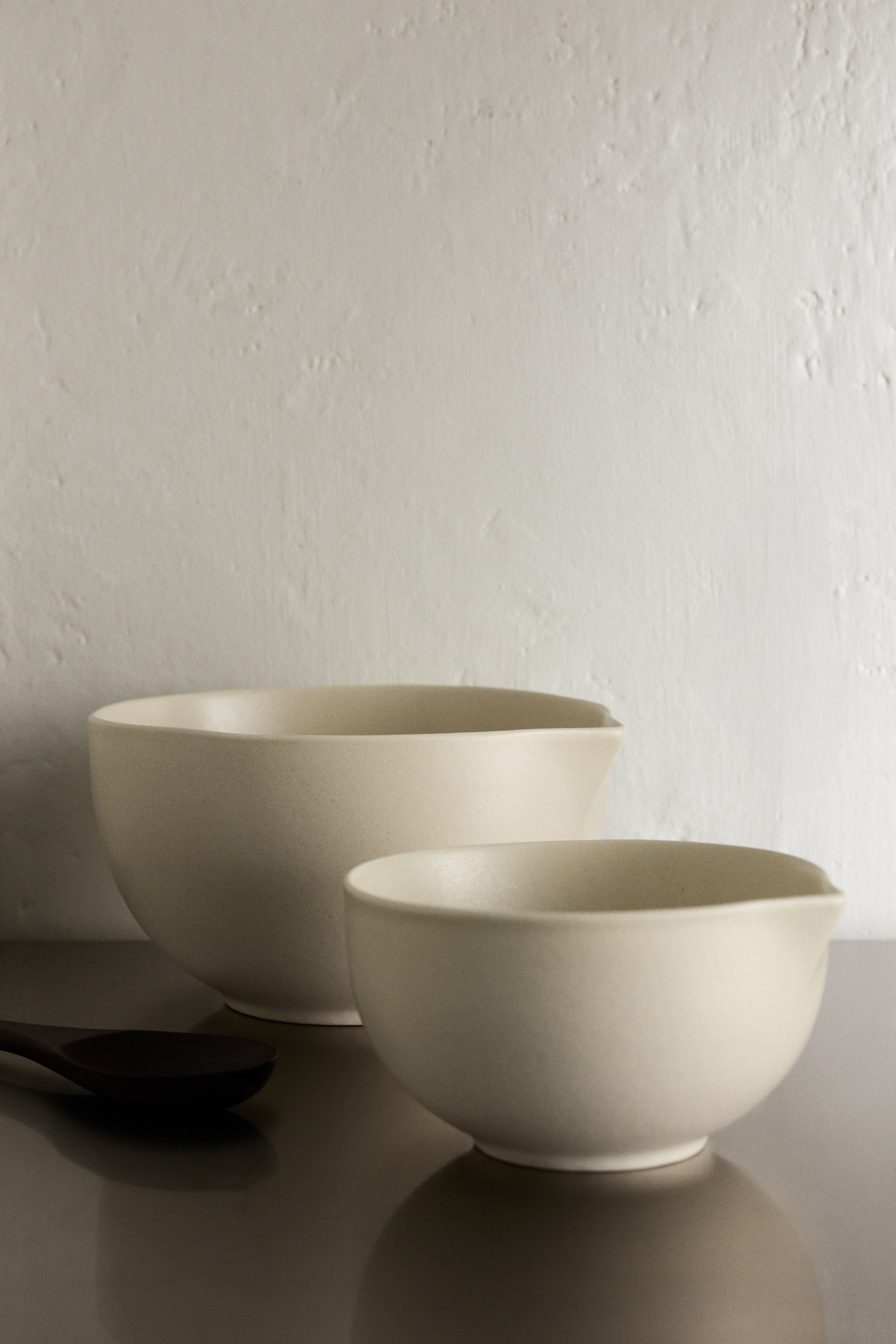 View larger image: Two cream-coloured ceramic bowls have a matt finish and each has a pouring spout on one side. The smaller bowl sits in front, angled slightly towards the viewer, with the larger bowl positioned behind it. A dark wooden spoon is resting, partially visible, on the surface in front.