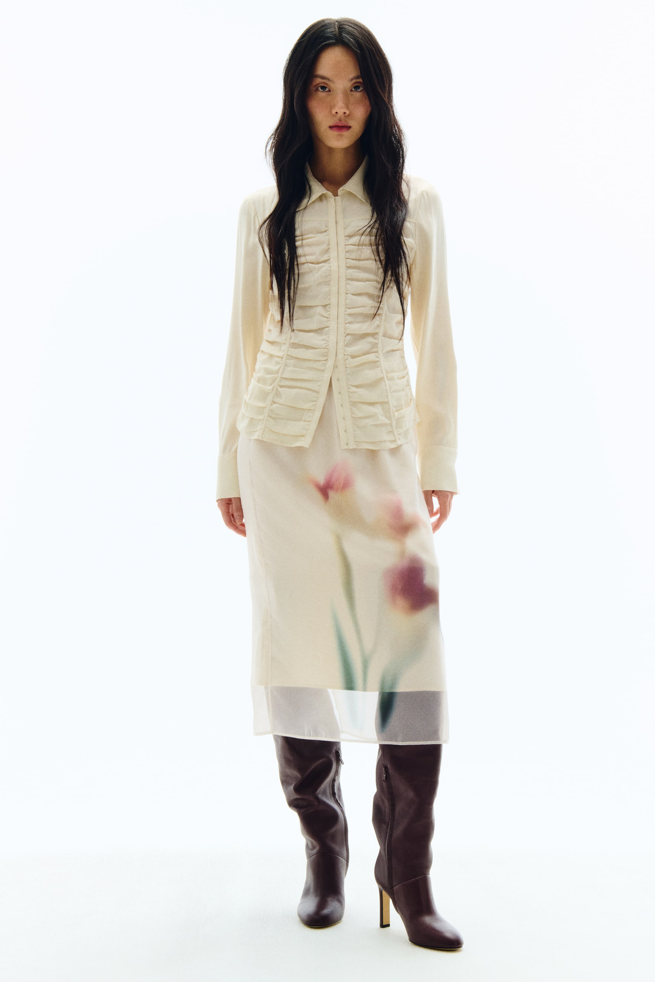 拡大画像を表示: クリームカラーの長袖シャツは、フロント中央にホック留めのギャザーパネルをあしらい、ぼかし調のフローラルプリントが施されたシアーなクリーム色のミディスカートと、ダークブラウンのニーハイヒールブーツを合わせたスタイリングです。