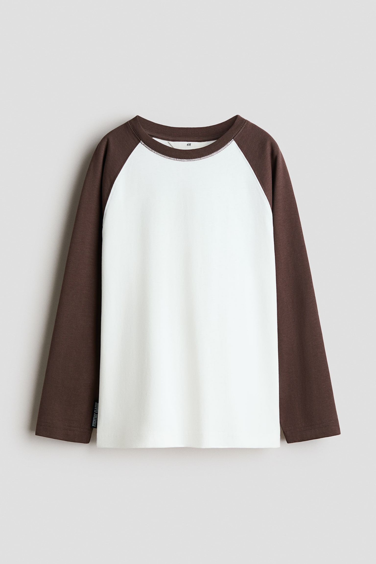 En lÃ¥ngÃ¤rmad raglant-shirt i loose fit visas framifrÃ¥n, med vit kropp och kontrasterande mÃ¶rkbruna lÃ¥nga Ã¤rmar. T-shirten har en matchande brun rund halsringning med ribbkant och en rak nederkant. - 1