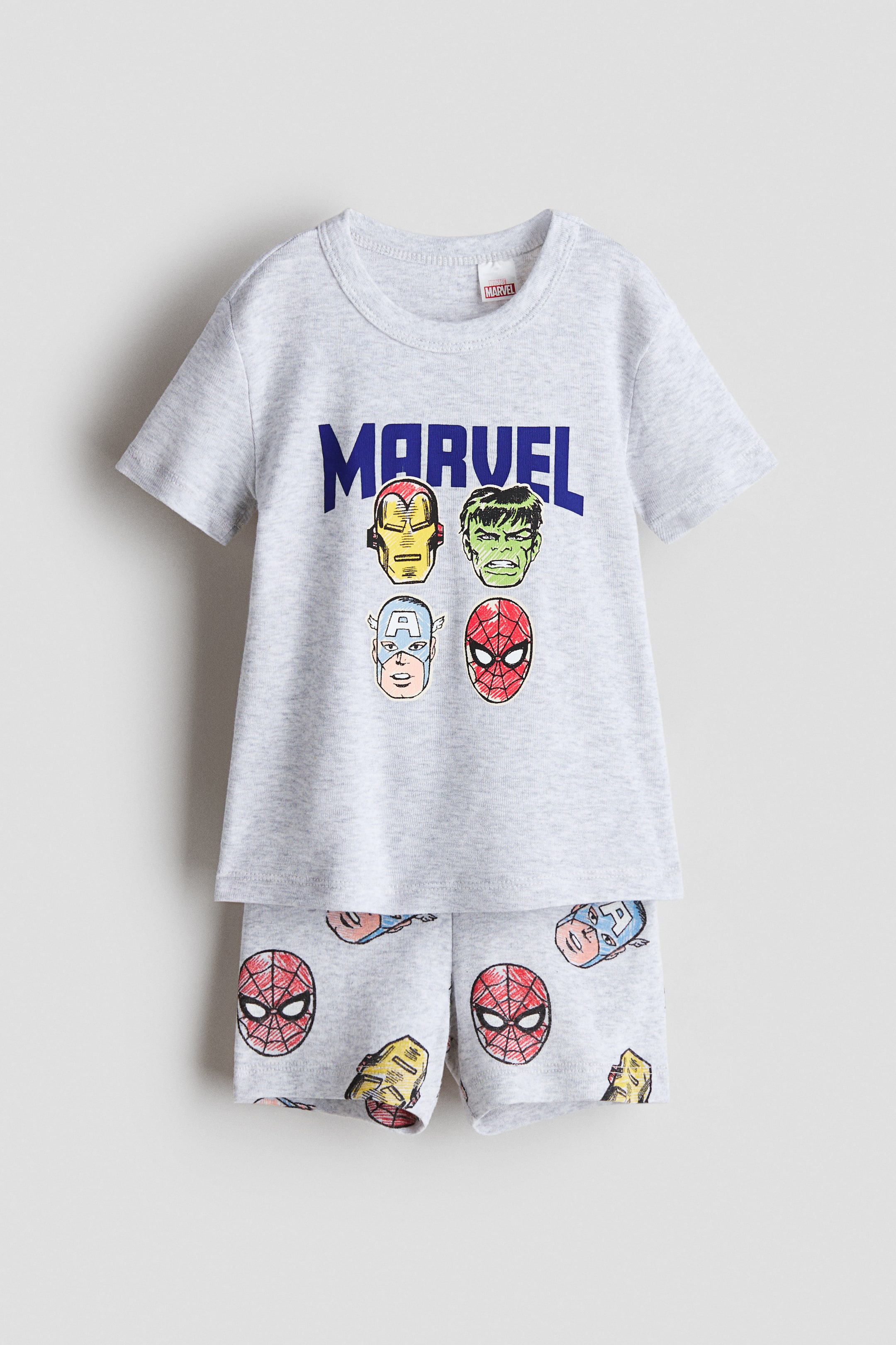 View larger image: A front-facing pajama set features a light gray short-sleeve T-shirt and matching shorts, both made from soft cotton jersey. The T-shirt displays blue 'MARVEL' text with four distinct cartoon character faces beneath it. The shorts have an elastic waistband and an all-over print of various cartoon character faces.