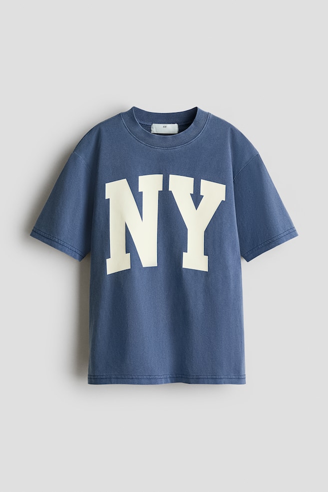 A loose-fit faded blue t-shirt is showcasing its front, featuring a large cream "NY" text printed across the chest. It is designed with a round, ribbed neckline and gently dropped shoulders. - 1