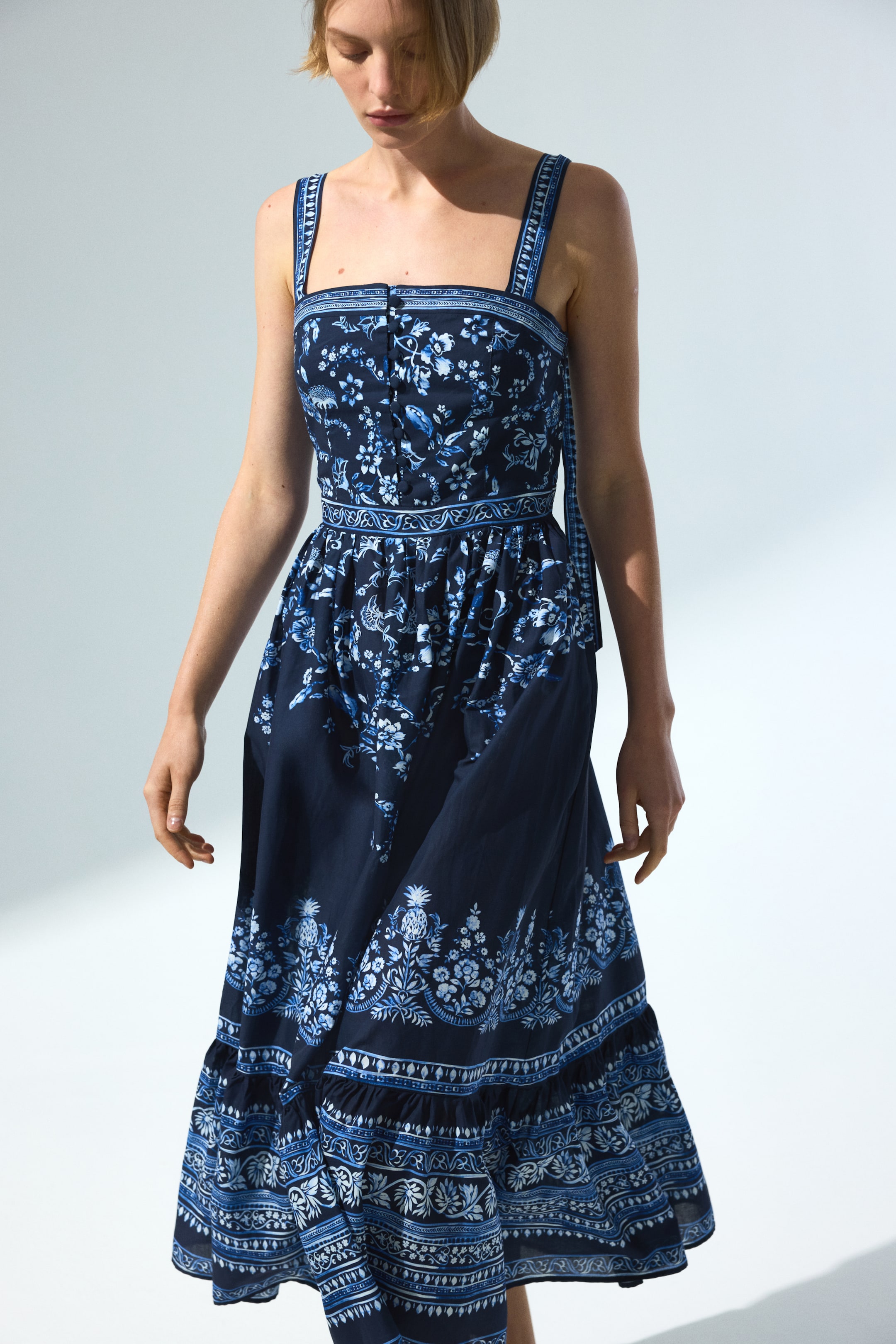 הצגת תמונה גדולה יותר: שמלת מידי בצבע כחול כהה, פונה מעט ימינה, מציגה דפוסים פרחוניים ואורנמנטליים מורכבים בצבעי כחול בהיר ולבן לאורך כל עיצוב השמלה. השמלה כוללת מפתח מרובע, רצועות כתף רחבות, חזית עם כפתורים, חגורת מותניים בדוגמה ייחודית, וחצאית מדורגת המסתיימת בשוליים מקומטים.