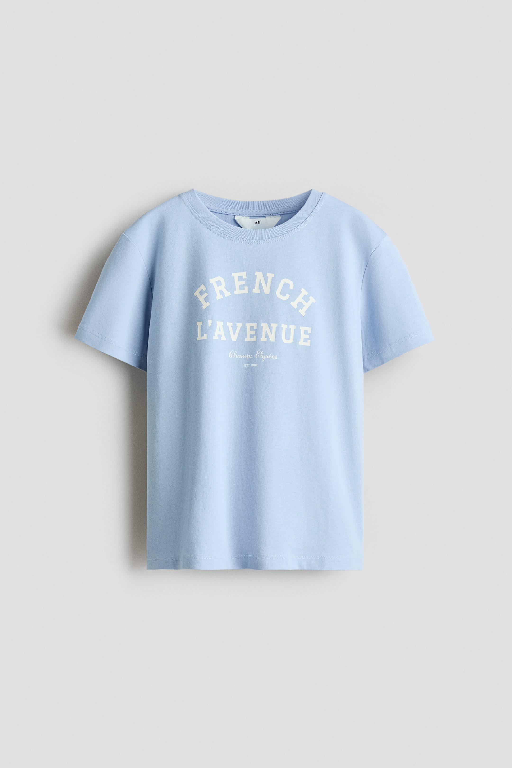 查看大圖: 淺藍色T恤以柔軟的棉質針織布製成,從正面展示,特色是前面的印花圖案。印花顯示「FRENCH L'AVENUE」以大而拱形的奶油色字母呈現,下方有較小的「Champs �lys�es」和「EST. 1997」字樣,T恤的圓領口則以羅紋邊飾收尾。