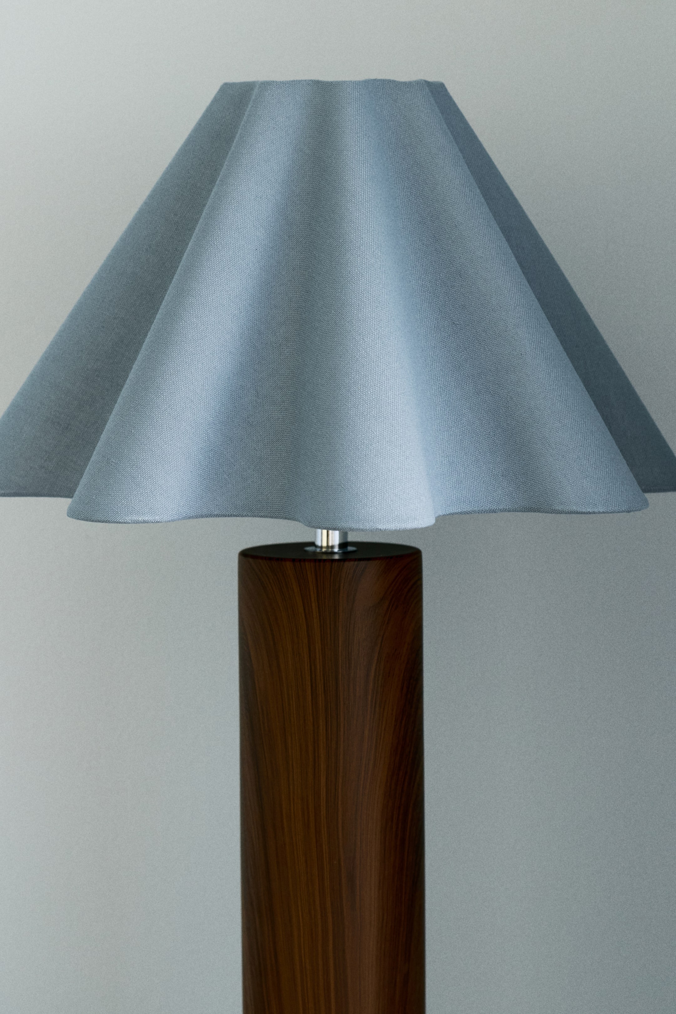 Grotere afbeelding bekijken: Een lamp met een donkerbruine cilindrische houten voet, die verbonden is met een lichtblauwe stoffen lampenkap met een zilverkleurig metalen onderdeel. De lampenkap vertoont zachte verticale plooien, waardoor een subtiel golvende onderrand ontstaat.