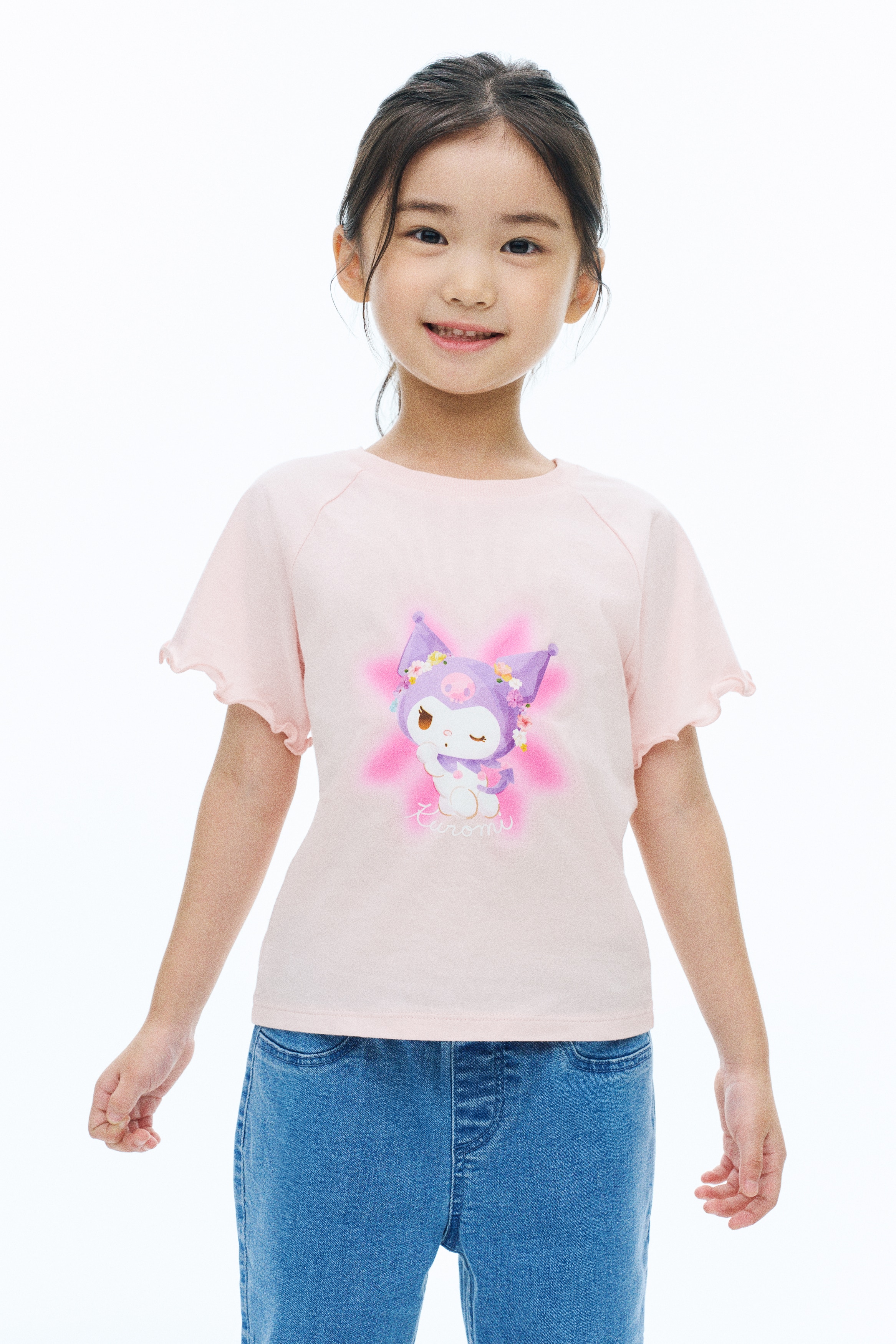 A light pink T-shirt is shown front-facing, featuring a ribbed crew neck and short raglan sleeves with ruffled lettuce-edge hems. The front of the shirt displays a central graphic of the character Kuromi wearing a floral crown, accented by a pink starburst background.
