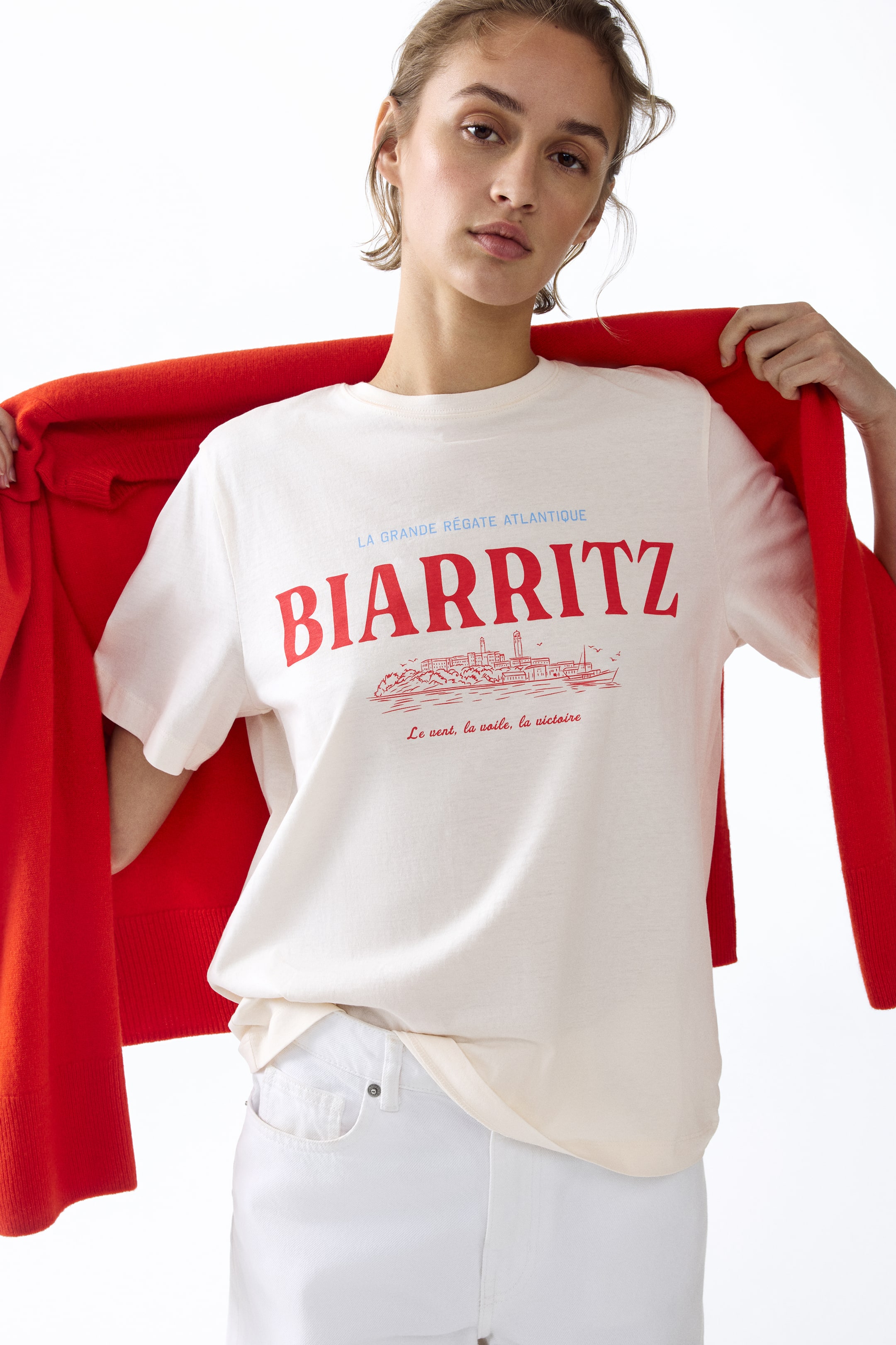 查看大圖: 一件米白色、短袖、圆领T恤，正面印有大红色“BIARRITZ”图案，搭配蓝色和红色的法文文字，以及一个红色轮廓的海滨小镇插图。T恤呈现出宽松的版型，衣摆自然垂落，肩上搭配一件红色开衫。