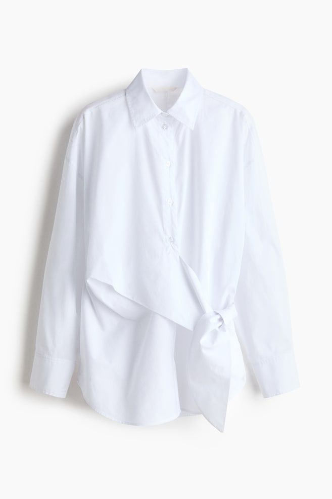 A white long-sleeved shirt is featuring a pointed collar and a front button placket, with a fabric panel wrapping from the left side and tying into a knot at the right hip. - 1