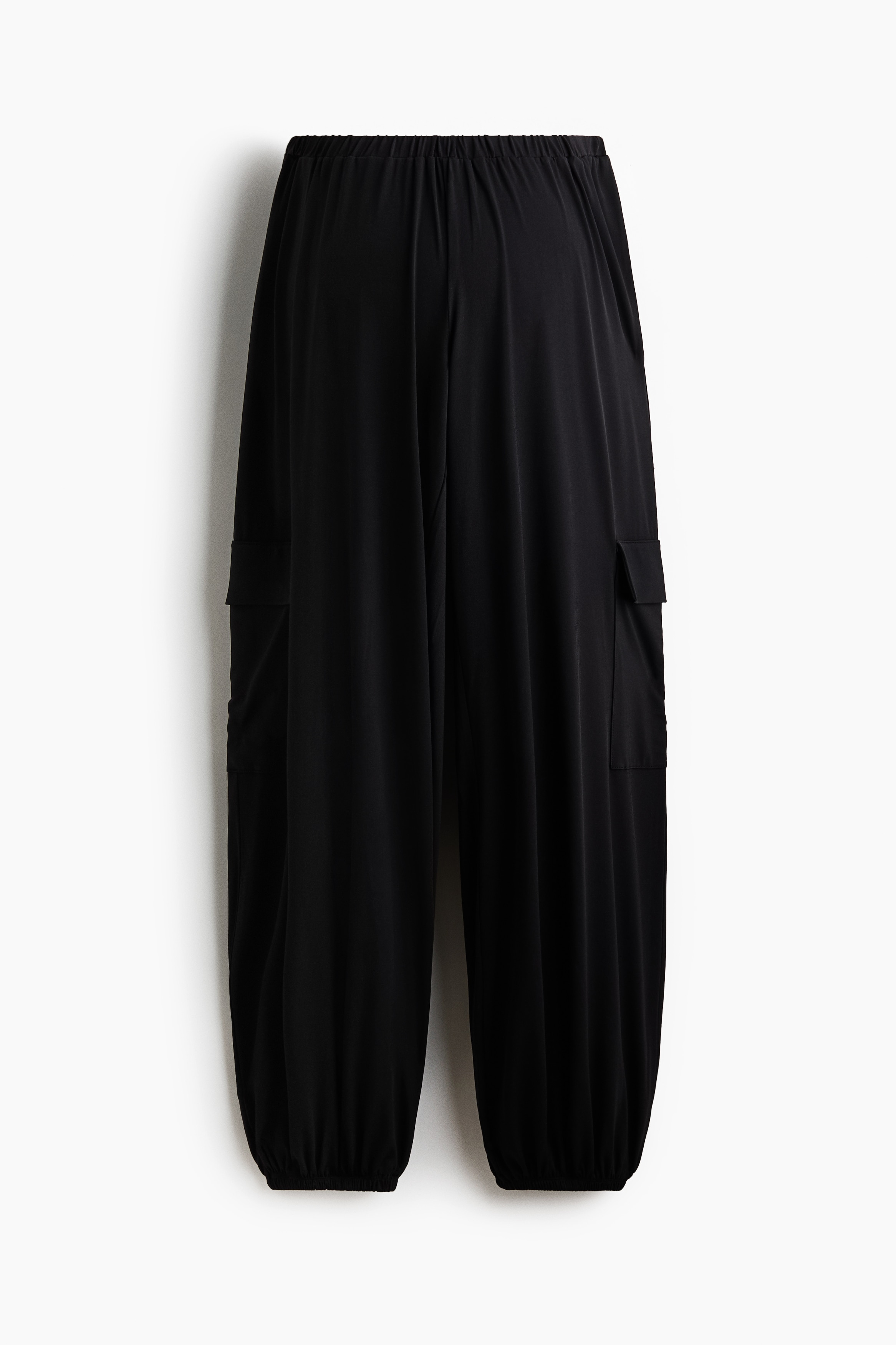 Pantalones balloon negros con cintura baja elástica y un corte holgado con caÃda, llevados por la modelo que está agachada. Los pantalones se combinan con un top crop a rayas blancas y negras y tenis negros.