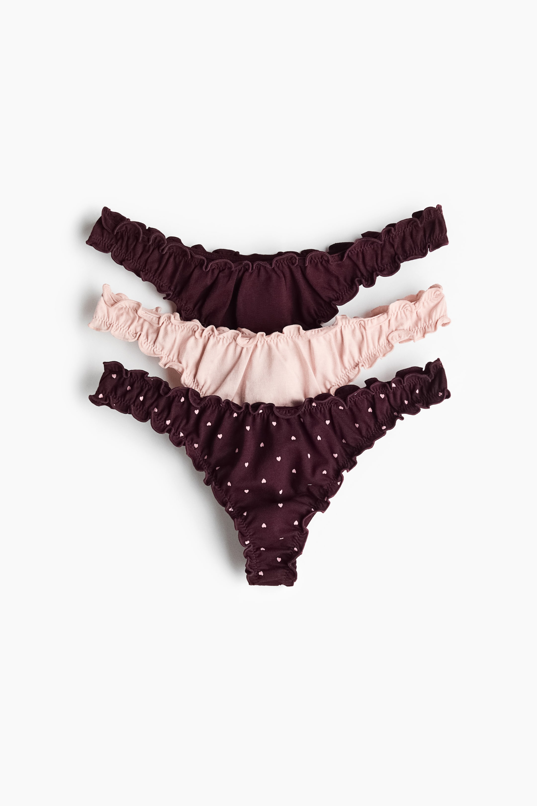 Grotere afbeelding bekijken: Drie stringslips worden weergegeven, met afgewerkte, gerimpelde randen en lage, V-vormige tailles. De set bevat een effen donker bordeauxkleurige slip, een effen lichtroze slip en een donker bordeauxkleurige slip met kleine lichtroze hartjespatronen.