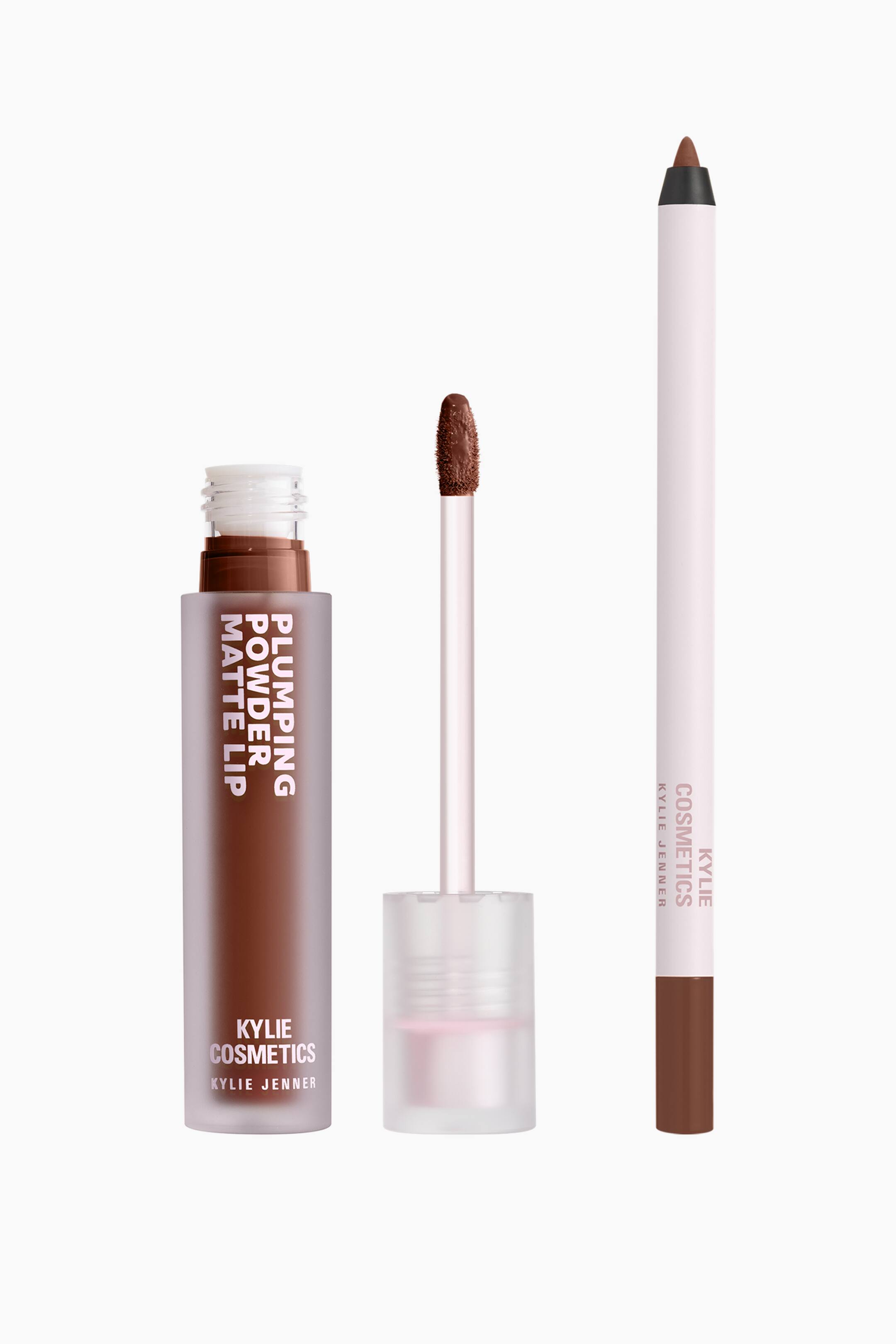 Visa större bild: Kylie Cosmetics Plumping Matte Lip Kit presenteras med tre komponenter i en brun nyans, placerade vertikalt sida vid sida mot en enfärgad bakgrund. Till vänster visas ett öppet, frostat rör med flytande läppstift och vit varumärkestext, intill en matchande applikatorstav med en doe-foot-spets i mitten, och till höger en vässad, ljusrosa läppennspenna med brun spets och ändhylsa.