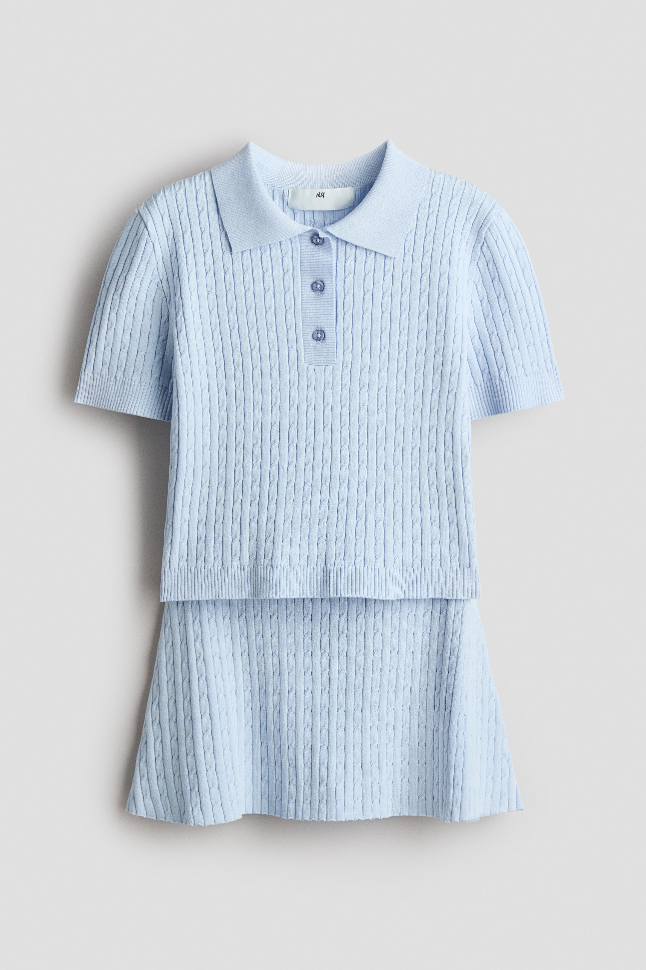 Agrandir l'image: Un ensemble en maille torsadée bleu clair est présenté de face, comprenant un polo à manches courtes et une jupe. Le polo est conçu avec un col, une patte de boutonnage à trois boutons et des finitions côtelées aux poignets et à l'ourlet, tandis que la jupe présente une taille élastiquée et un léger évasement à l'ourlet.