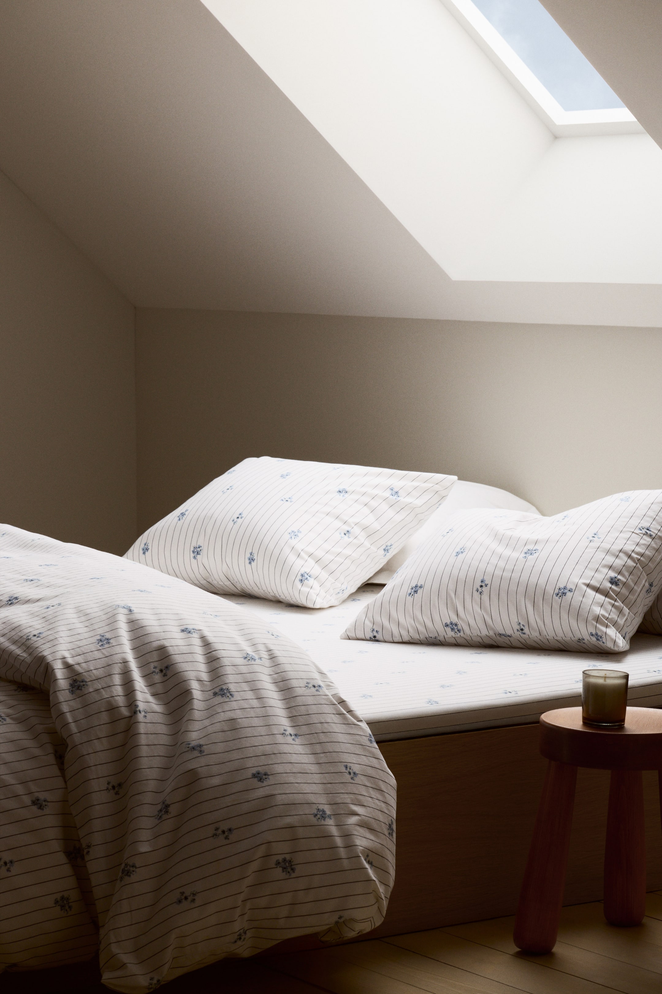 Преглед на по-голямо изображение: Комплект спално бельо е с бяла материя с тънки хоризонтални сини ивици и малки сини цветни клъстери. Комплектът включва одеало, покривало със зашити гънки и две калъфки за възглавници, всички показващи този координиран модел. Одеялото е поставено на леглото, а двете калъфки за възглавници са поставени.