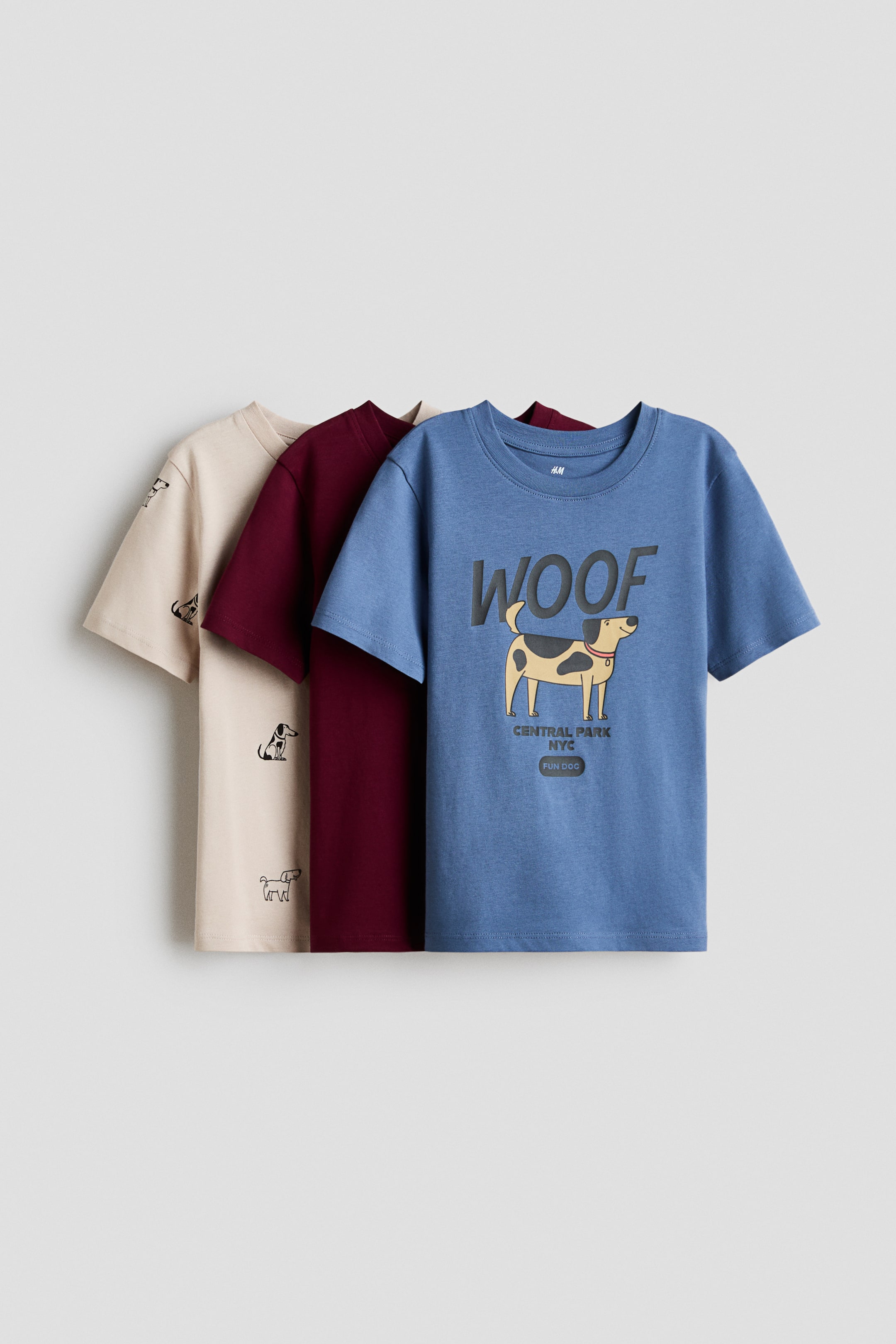 View larger image: Tatlong malalambot na cotton jersey T-shirts ang ipinapakita sa isang offset, layered na pagkakaayos, bahagyang nakatagilid sa kaliwa, bawat isa ay may round, rib-trimmed na neckline. Ang harapang asul na T-shirt ay may cartoon na graphic ng aso na may 'WOOF' at 'CENTRAL PARK NYC', ang gitnang maroon na T-shirt ay nagpapakita ng simpleng harapan, at ang likod na beige na T-shirt ay may maraming maliliit na itim na outline ng aso.