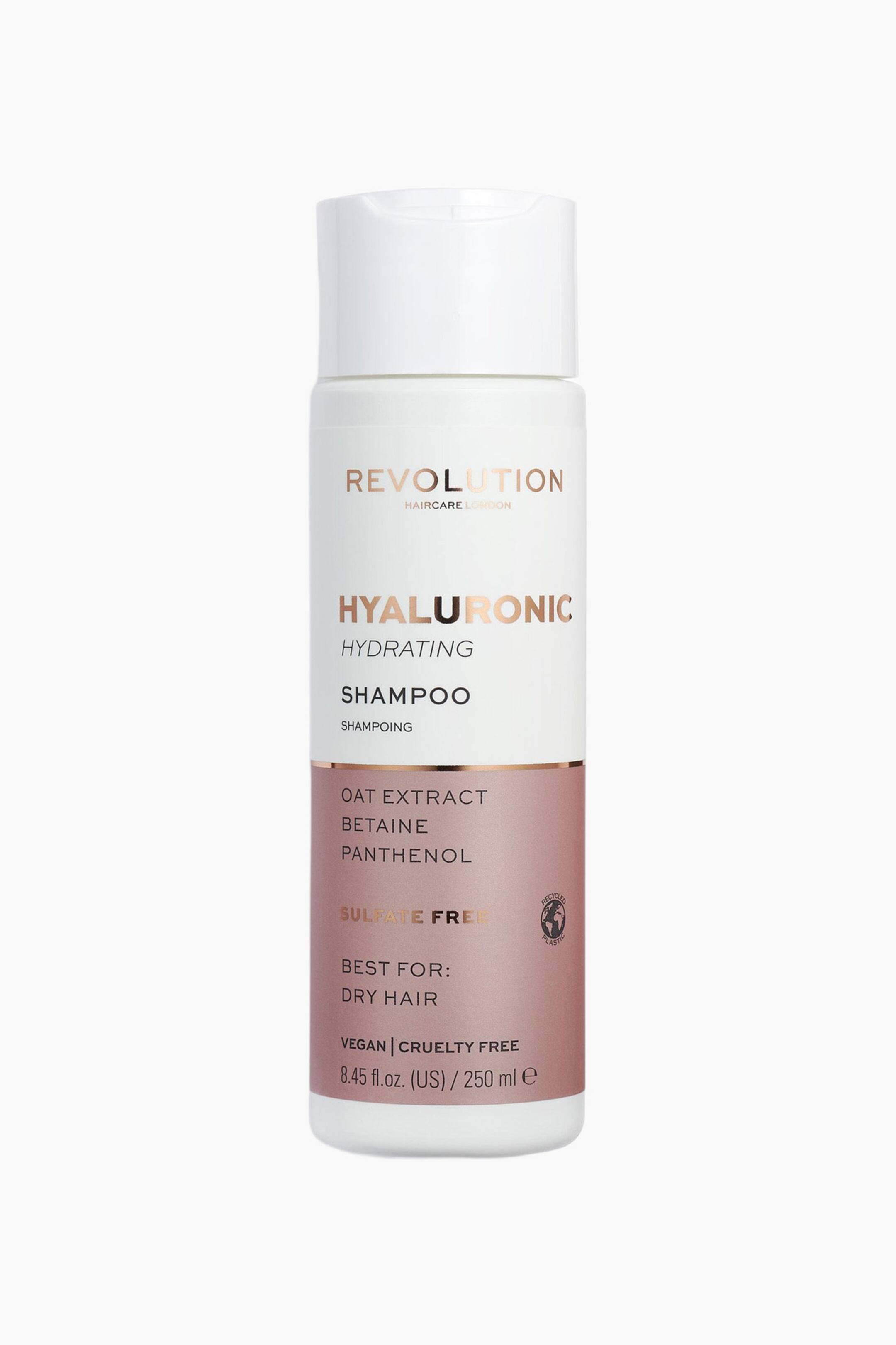 Se større billede: En cylindrisk flaske med Revolution Haircare Hyaluronic Hydrating Shampoo står oprejst og har en hvid flip-top-kapsel samt en frontvendt etiket. Etiketten viser rosaguld metallisk tekst på en hvid øverste del og en støvet rosa nederste blok, der oplister nøgleingredienser og produktoplysninger.