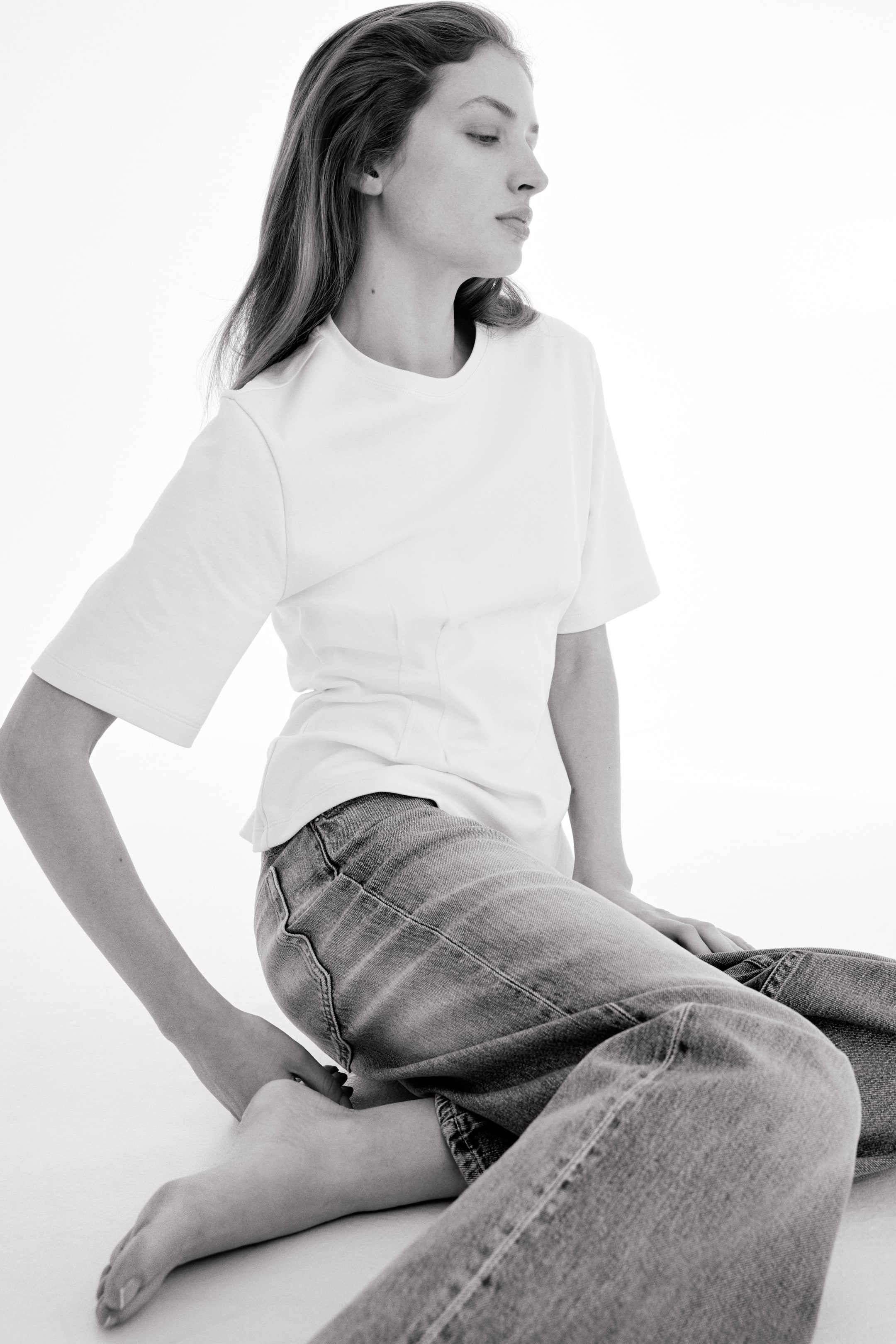 Agrandir l'image: Un haut blanc à manches courtes présentant une encolure ronde et des coutures verticales qui cintrent la taille, porté avec un jean bleu clair au délavage léger offrant une coupe décontractée, vu de trois-quarts.