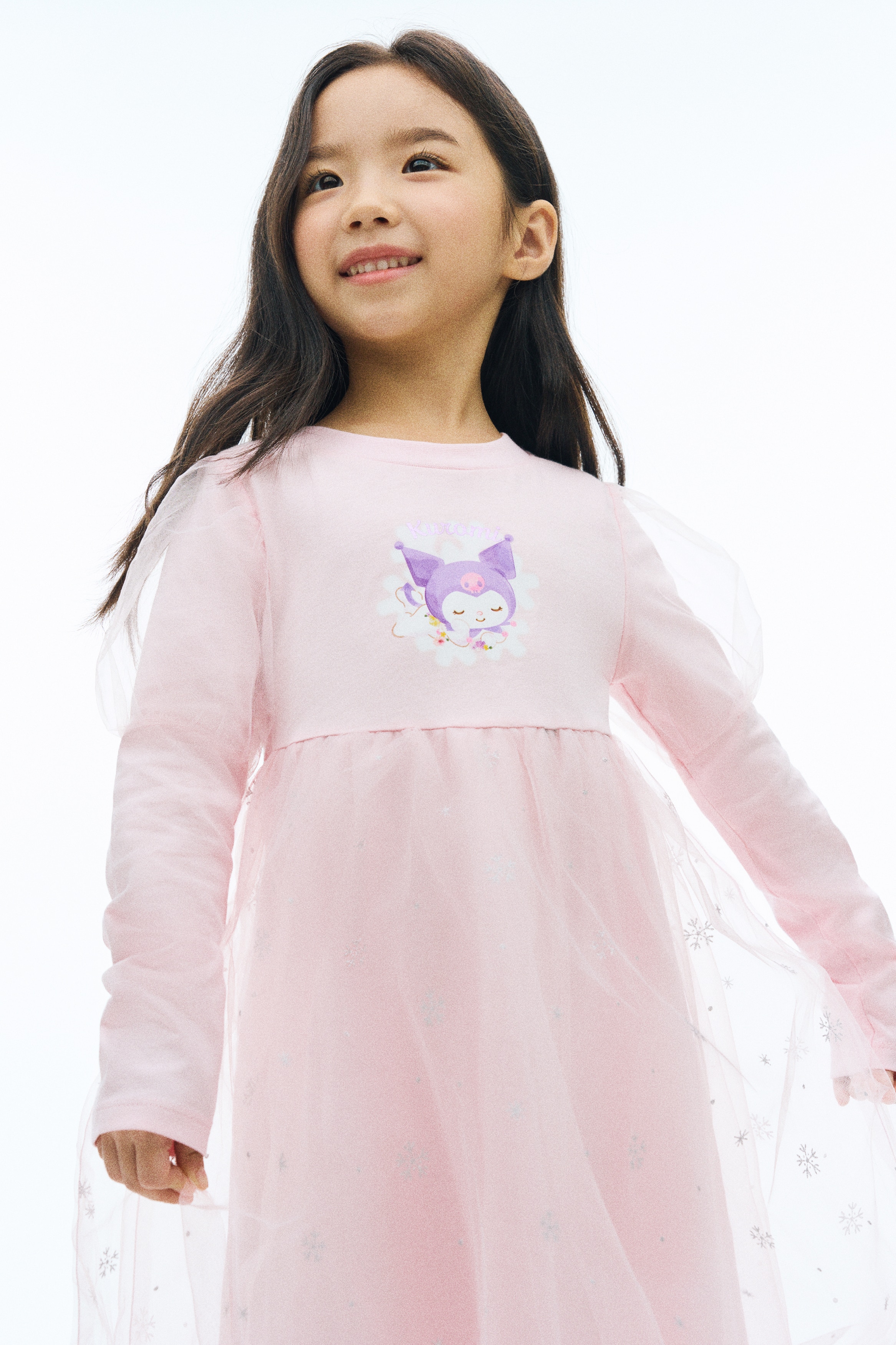 A light pink long-sleeved dress is shown from the front, featuring a round neckline and a Kuromi graphic on the chest. The dress features a gathered skirt with a sheer tulle overlay speckled with silver glitter snowflakes, and sheer tulle detailing at the shoulders.