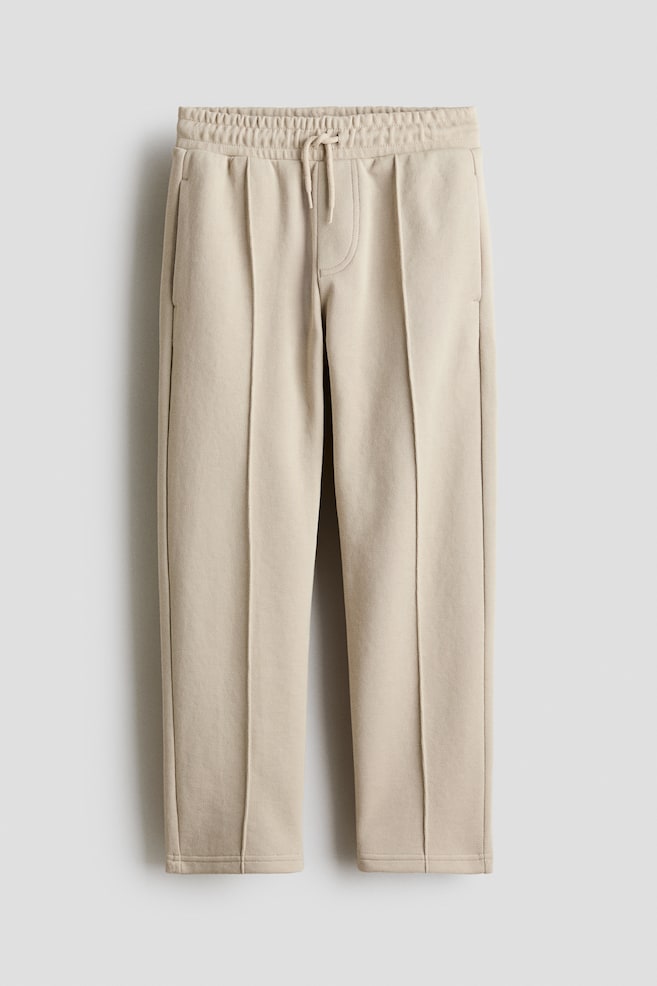 Los pantalones beige se muestran en vista frontal, con pretina elástica con cordón anudado, pliegues verticales cosidos a lo largo del frente de cada pierna, bolsillos laterales visibles y puños con dobladillo limpio. - 1