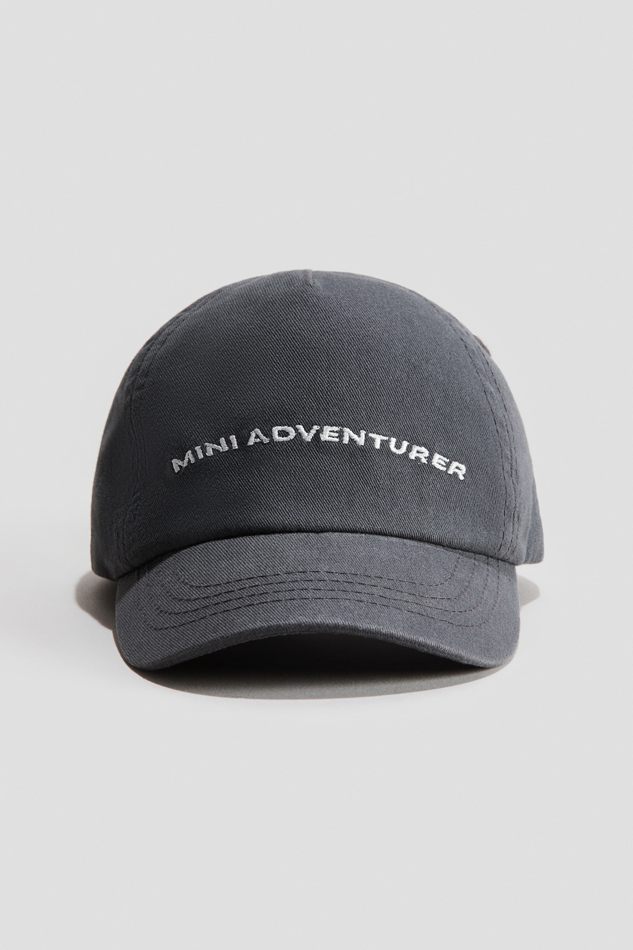 View larger image: The front view of a dark grey cap is displayed, showcasing "MINI ADVENTURER" embroidered in white text across the front panel. Crafted from a cotton weave fabric, the cap's brim features multiple rows of stitching.
