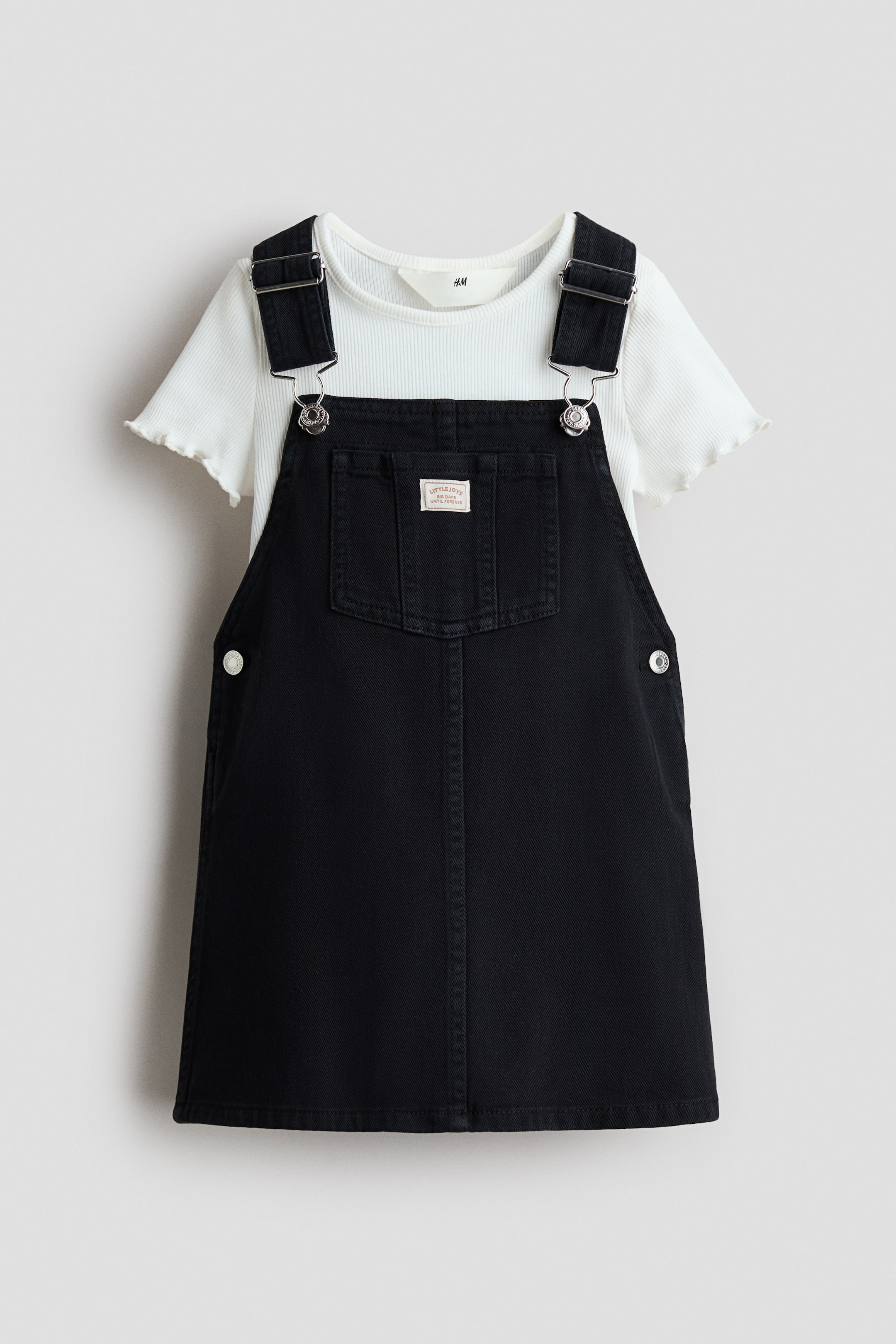 Grösseres Bild ansehen: Ein schwarzes Baumwoll-Twill-Latzkleid wird über einem weißen, gerippten Baumwolljersey-T-Shirt getragen, beide in FRONT-Ansicht. Das Latzkleid verfügt über verstellbare Schulterträger mit Metallverschlüssen, eine aufgesetzte Brusttasche mit einem kleinen Etikett und Druckknöpfe an den Seiten, während das T-Shirt einen schmalen Besatz um den Halsausschnitt und überlockte, geraffte Kanten an den kurzen Ärmeln aufweist.
