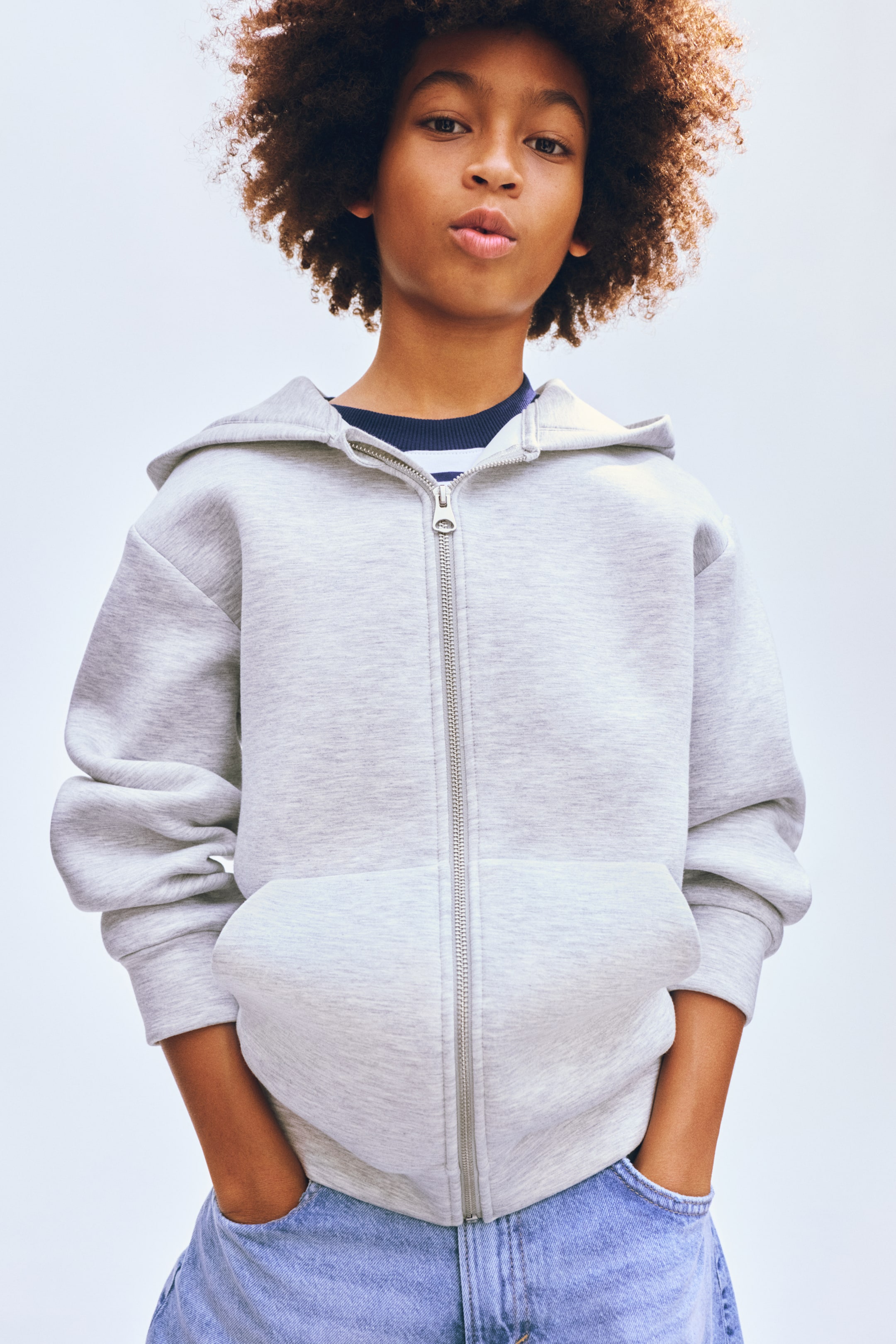 View larger image: The light grey full-zip hoodie showcases a silver-toned zipper running down the centre front, a structured hood, and a large kangaroo pocket with hands resting inside. It also features ribbed cuffs and a ribbed hem, and is worn over a navy and white striped top.