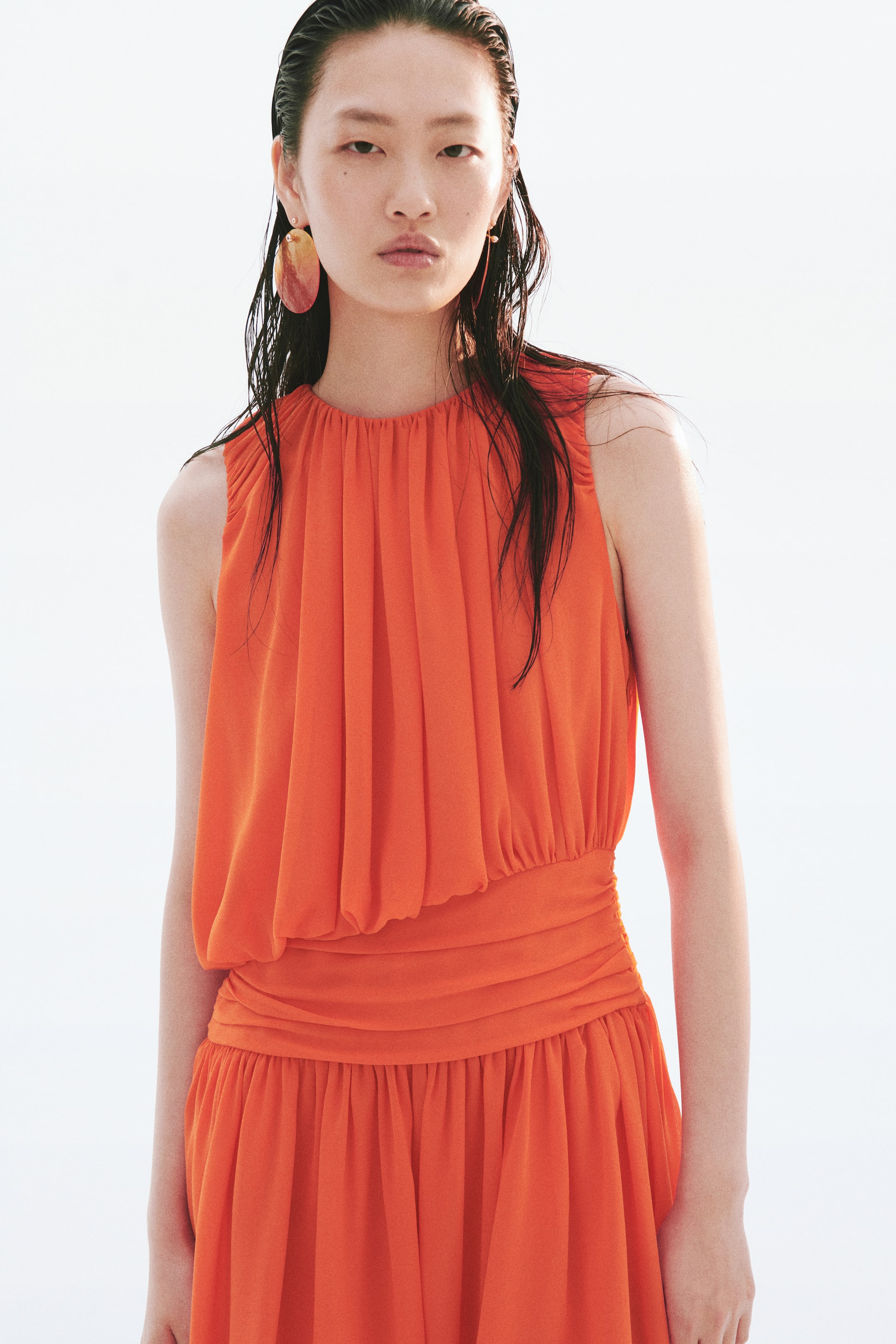 View larger image: An orange sleeveless dress featuring a gathered neckline and a loose bodice, cinched by a wide ruched waistline. The gathered skirt flows from the waist, with the dress shown from a slight front-right angle.