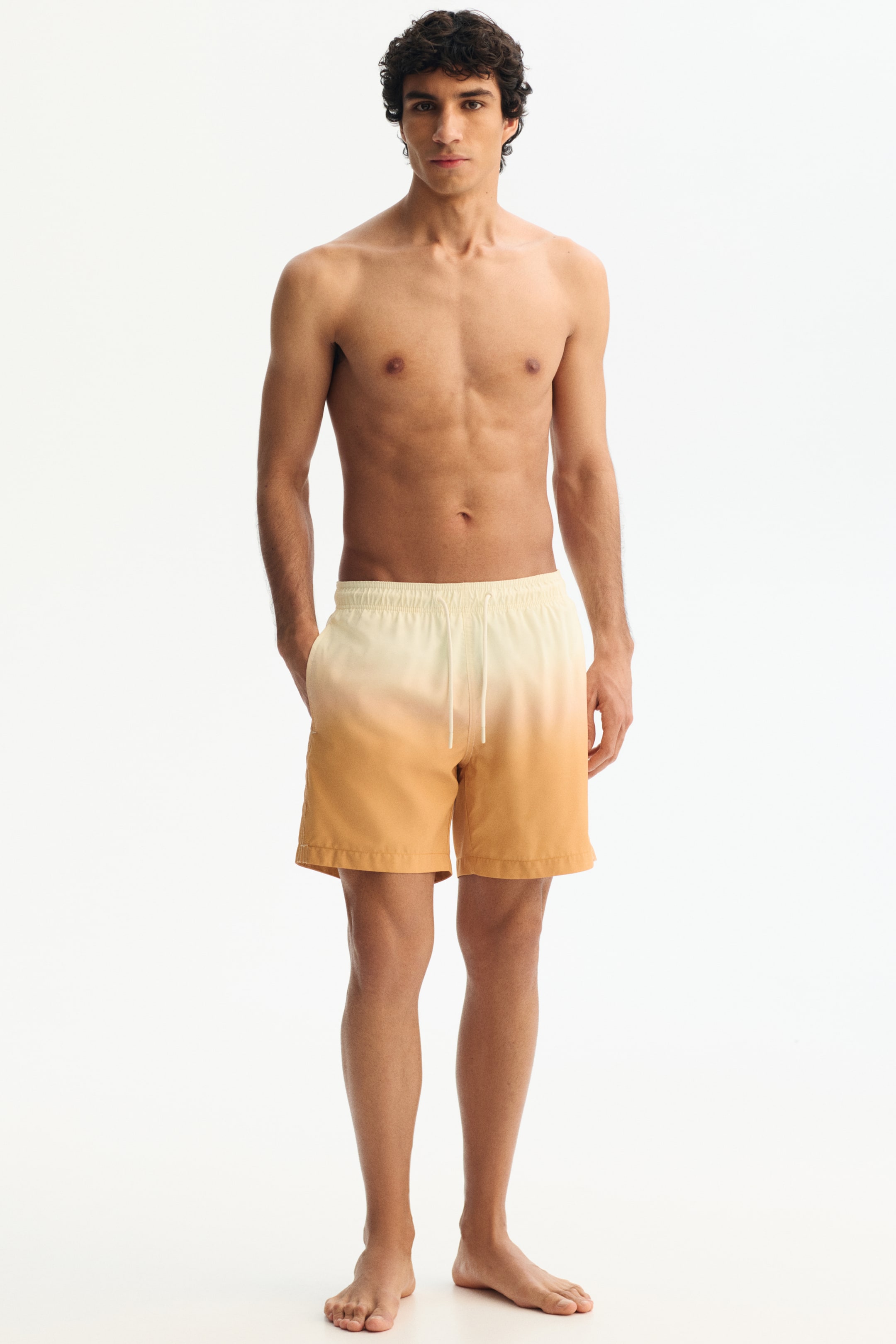 View larger image: Men’s swim shorts showcase an ombre pattern, shifting from light cream at the waist to golden yellow at the hem, as seen from a front view. They feature an elastic waistband with a white drawstring and side pockets, extending to a mid-thigh length.