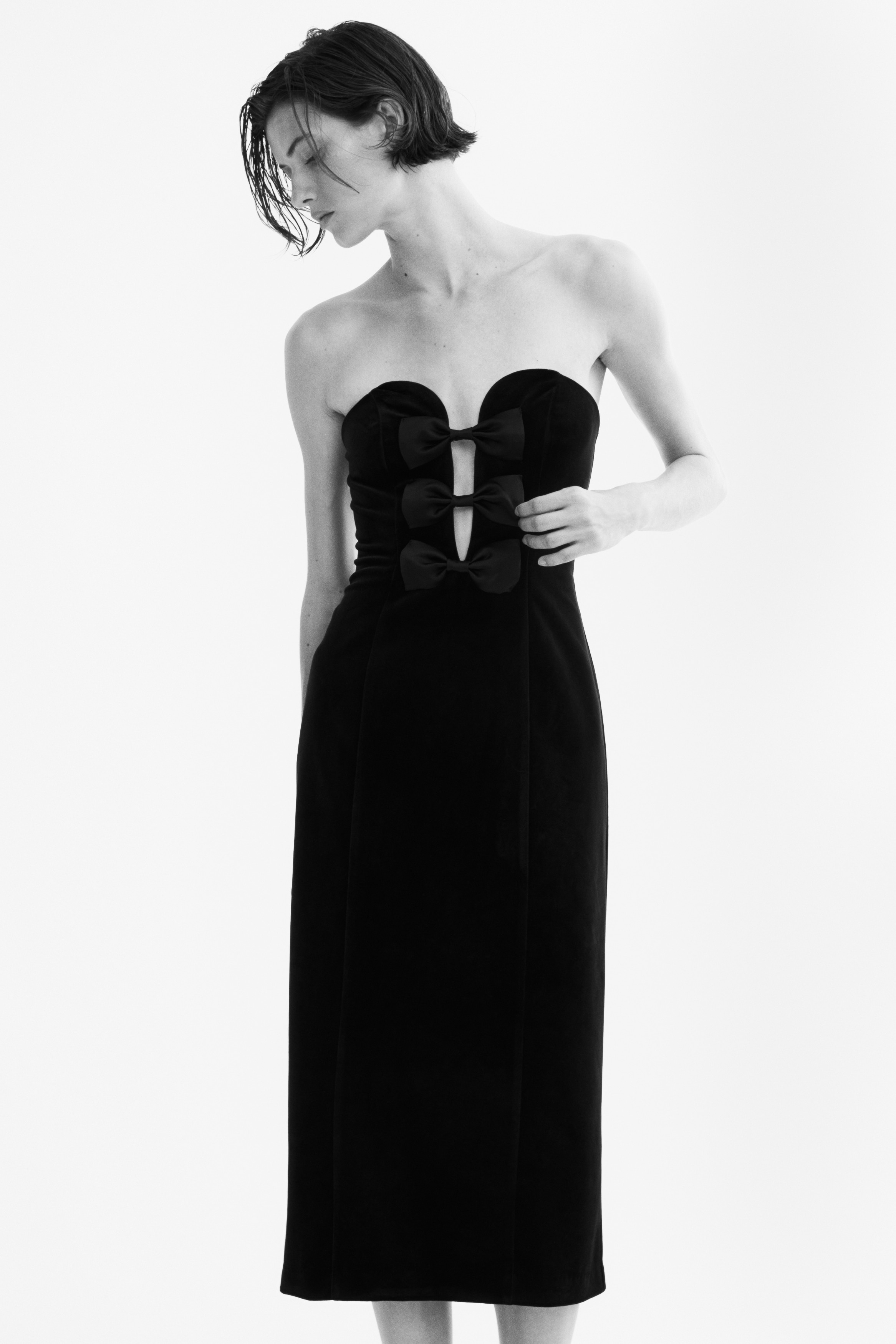 A black, calf-length bandeau dress is shown from the front, featuring a structured sweetheart neckline and three decorative black satin bows stacked vertically down the center of the bodice. The dress is crafted from a soft, matte velour fabric and has a fitted silhouette.