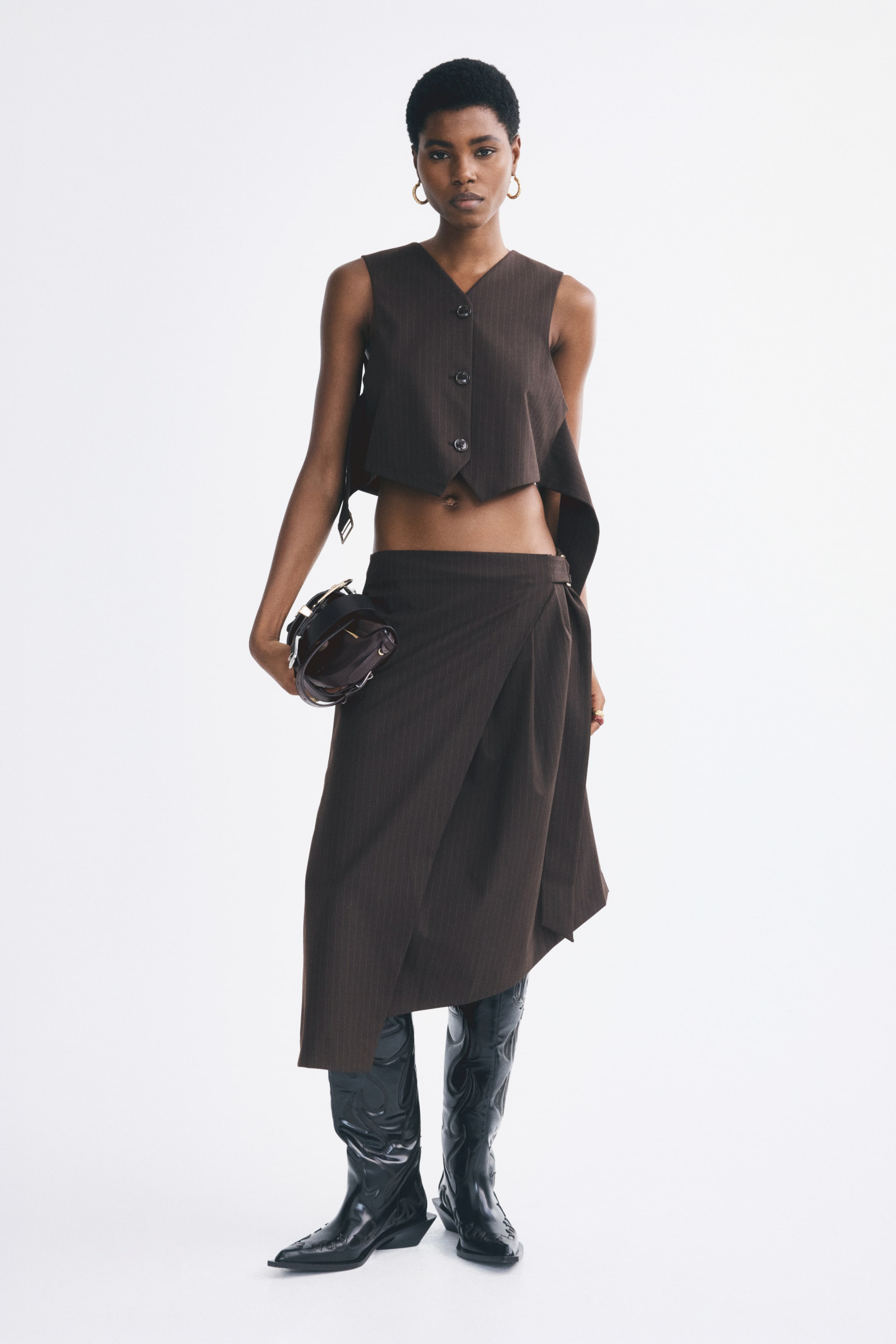 View larger image: The model is wearing a cropped, fitted vest in dark brown pinstriped fabric, featuring a V-neckline, narrow shoulder cut, and three front buttons. The vest is styled with a matching wrap-style midi skirt and black knee-high boots, while a small dark handbag is held in one hand.