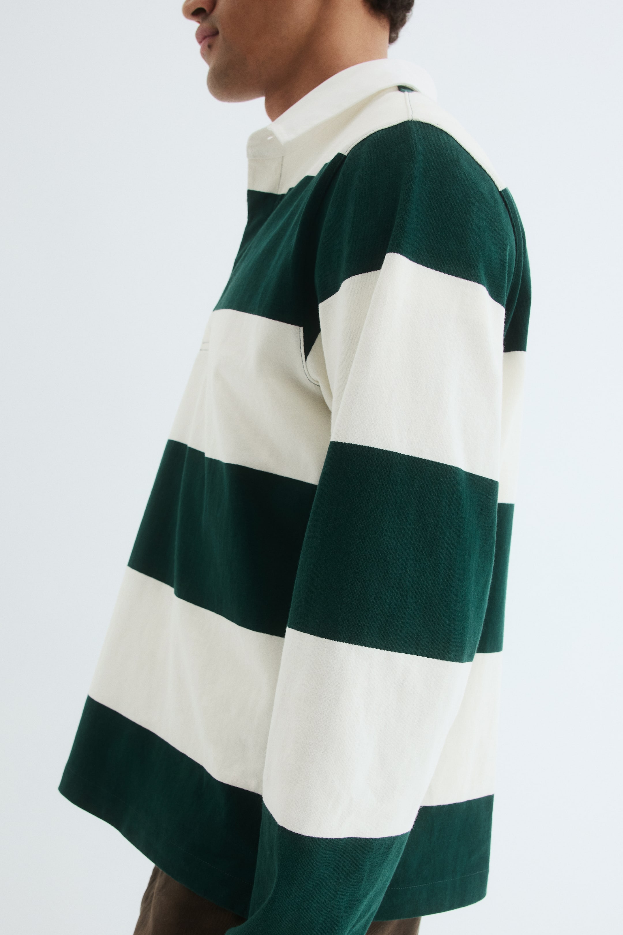 View larger image: A striped rugby shirt is being worn, angled to the left side, featuring wide horizontal stripes in dark green and off-white. It has a contrasting white collar and long sleeves, with the fabric draping loosely.