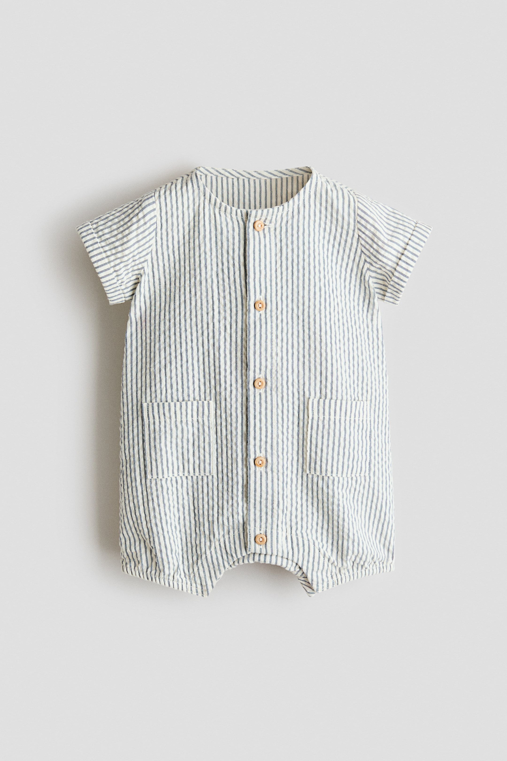 View larger image: A romper suit in a narrow vertical stripe pattern is displayed at the front, featuring a round neckline and a full button-down placket with wooden-look buttons. It also showcases short sleeves with fixed turn-up cuffs, two front patch pockets, and elasticated leg hems.