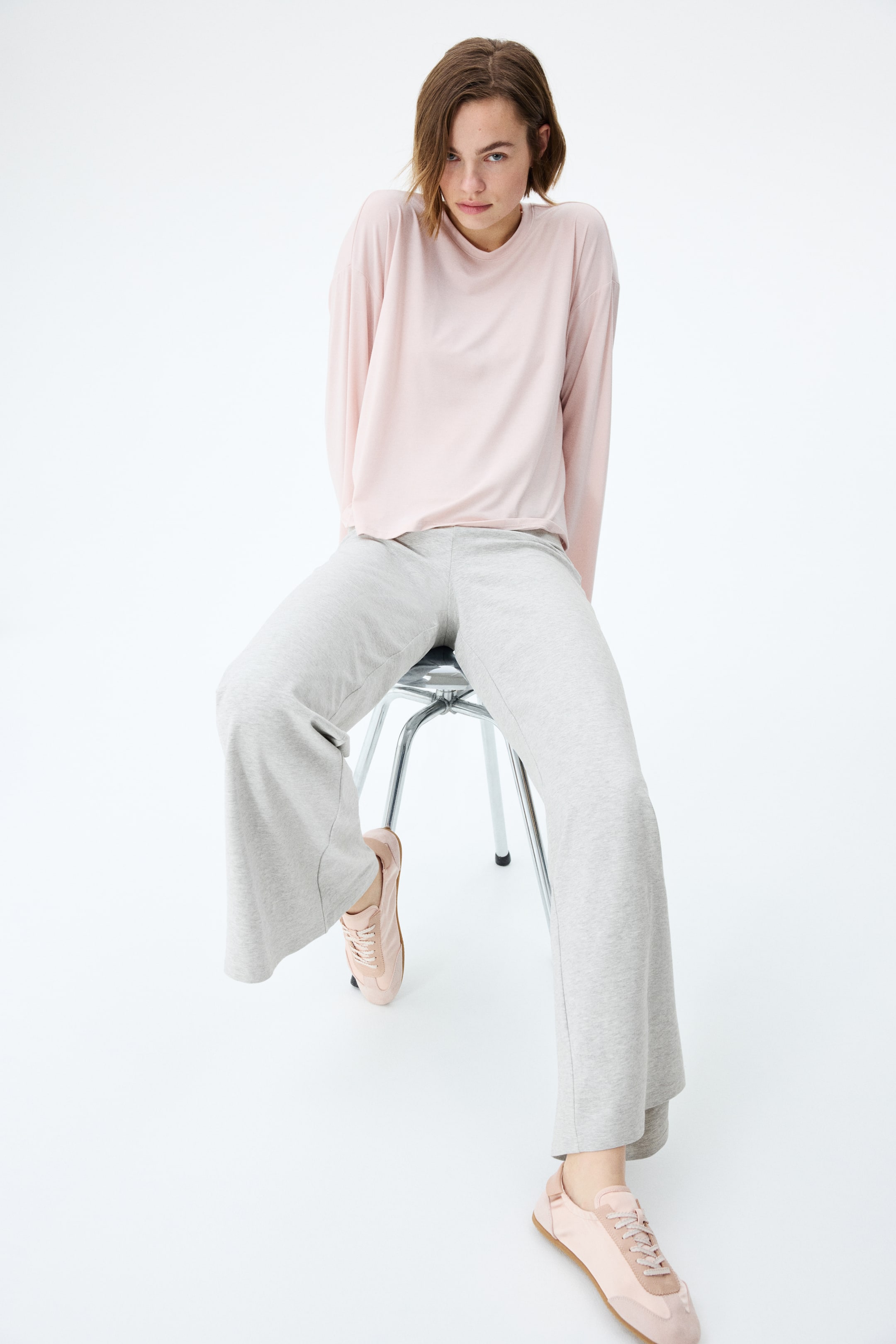 View larger image: A blush pink long-sleeve top with a crew neck drapes loosely, paired with light grey wide-leg pants that flow, showcasing their full, relaxed cut. Light pink lace-up sneakers complete the look.