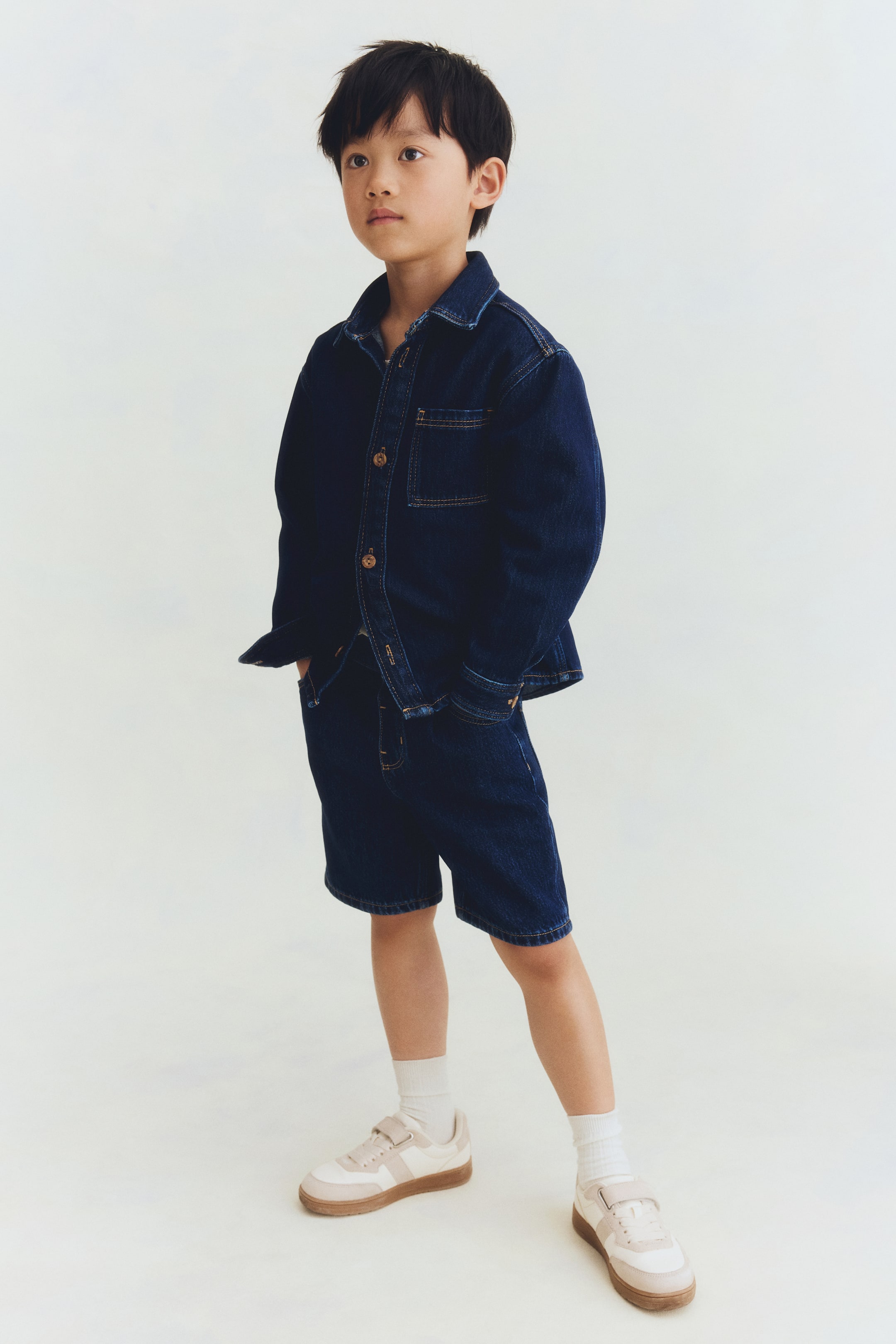 Visa större bild: En mörk tvättad denimskjorta och matchande shorts presenteras från en lätt frontal vinkel. Skjortan har en krage, frontknappar och en enkel bröstficka med kontrastsömmar, medan shortsen sträcker sig till knälängd.