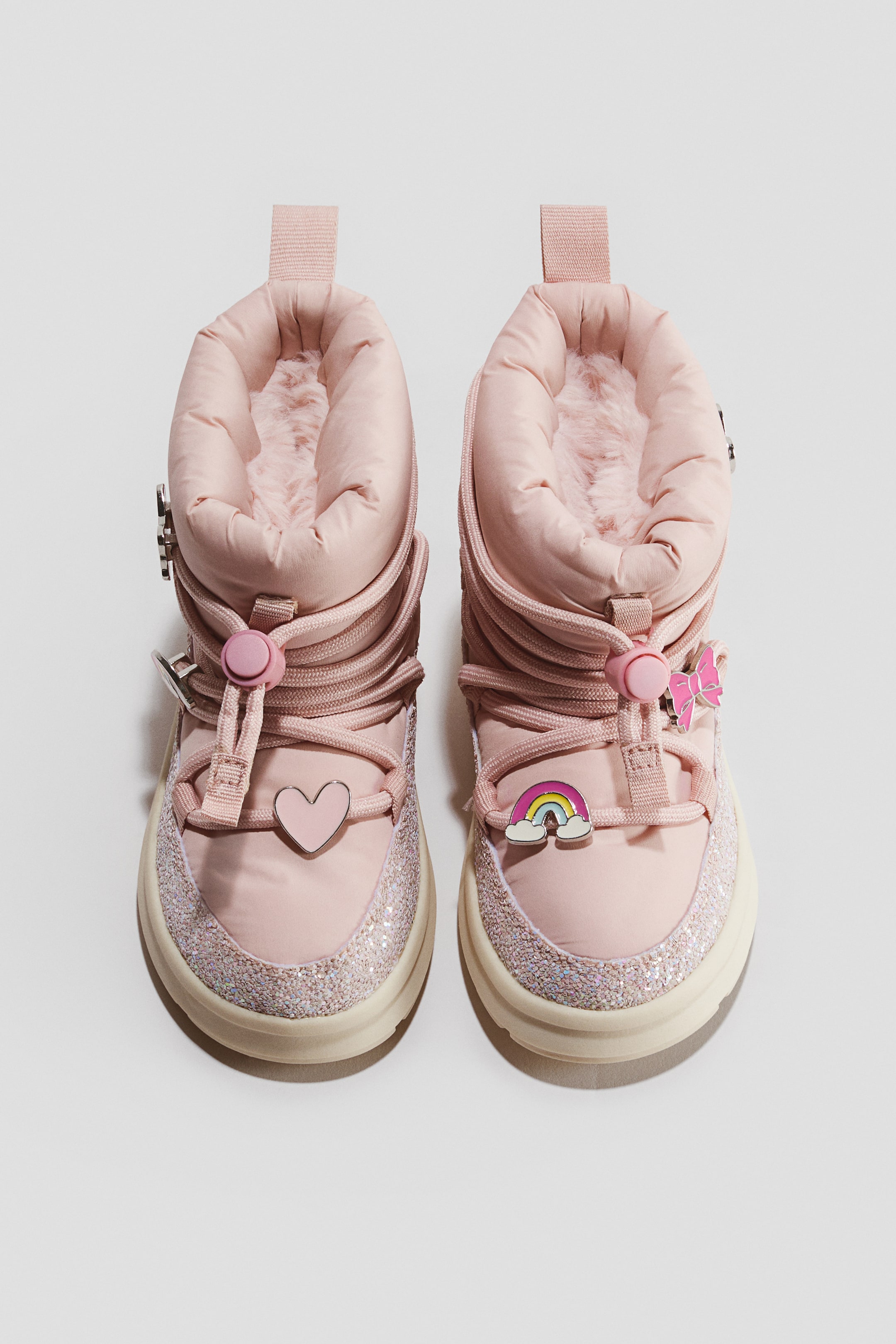 Agrandir l'image: Une paire de bottes d’hiver bouffantes rose clair est présentée de face, avec un col de cheville rembourré, une doublure douce en fausse fourrure et des lacets en cordon entourés d’un système de fermeture à stoppeur. Les bottes présentent des breloques décoratives, notamment un cœur et un arc-en-ciel sur le bout de la chaussure, et sont conçues avec une bordure scintillante le long du garde-boue ainsi qu’une semelle en caoutchouc couleur crème.