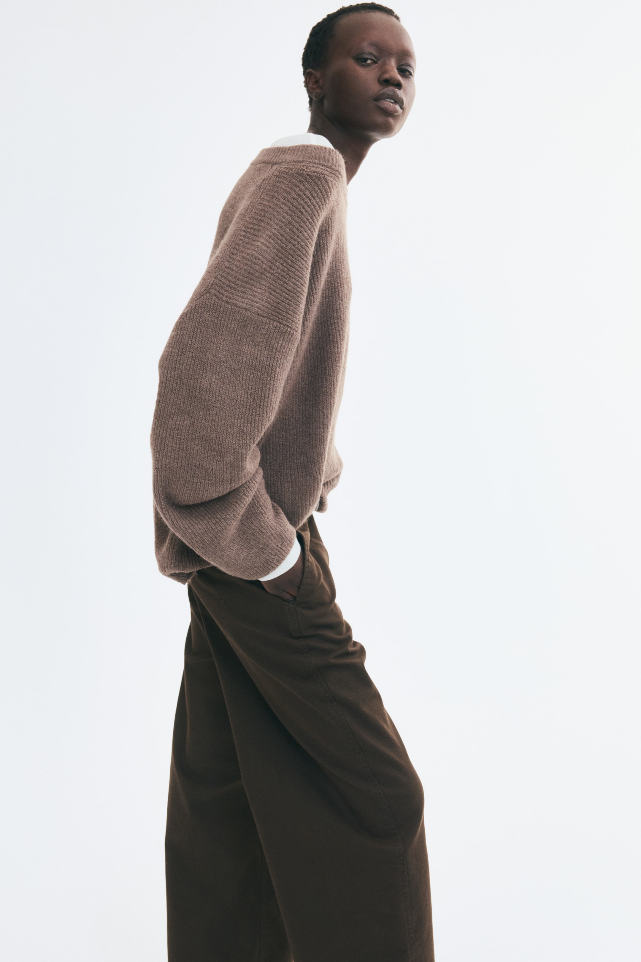 View larger image: The model is wearing a taupe ribbed-knit sweater with dropped shoulders and a relaxed fit. The sweater is layered over a white shirt with visible cuffs and collar and paired with dark brown trousers.