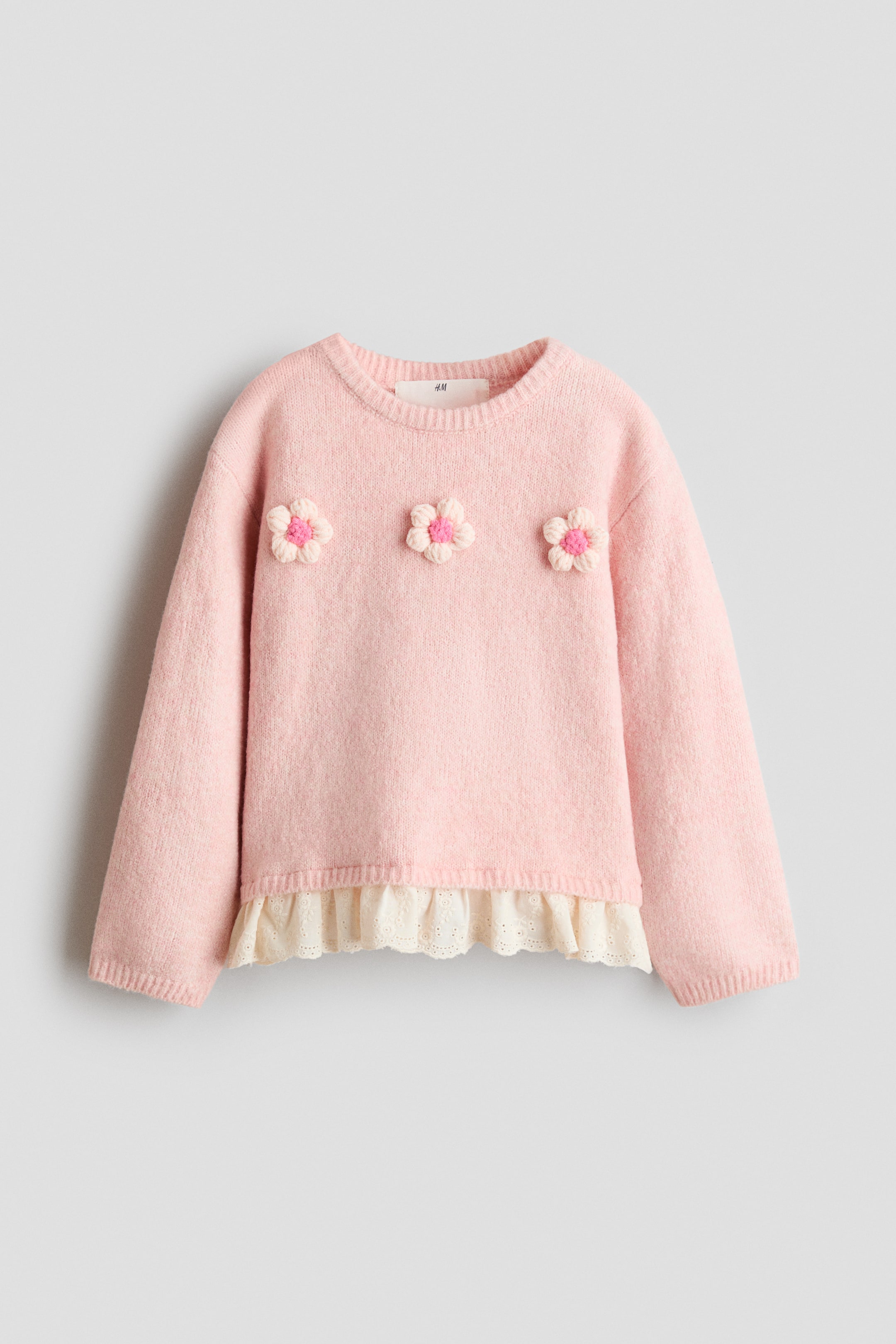 View larger image: A light pink knit sweater is shown front-facing, featuring a ribbed crew neck, long sleeves, and three crocheted 3D flowers with pink centres arranged across the chest. A white, ruffled eyelet-embroidered hem extends from beneath the bottom hem, creating a layered appearance.
