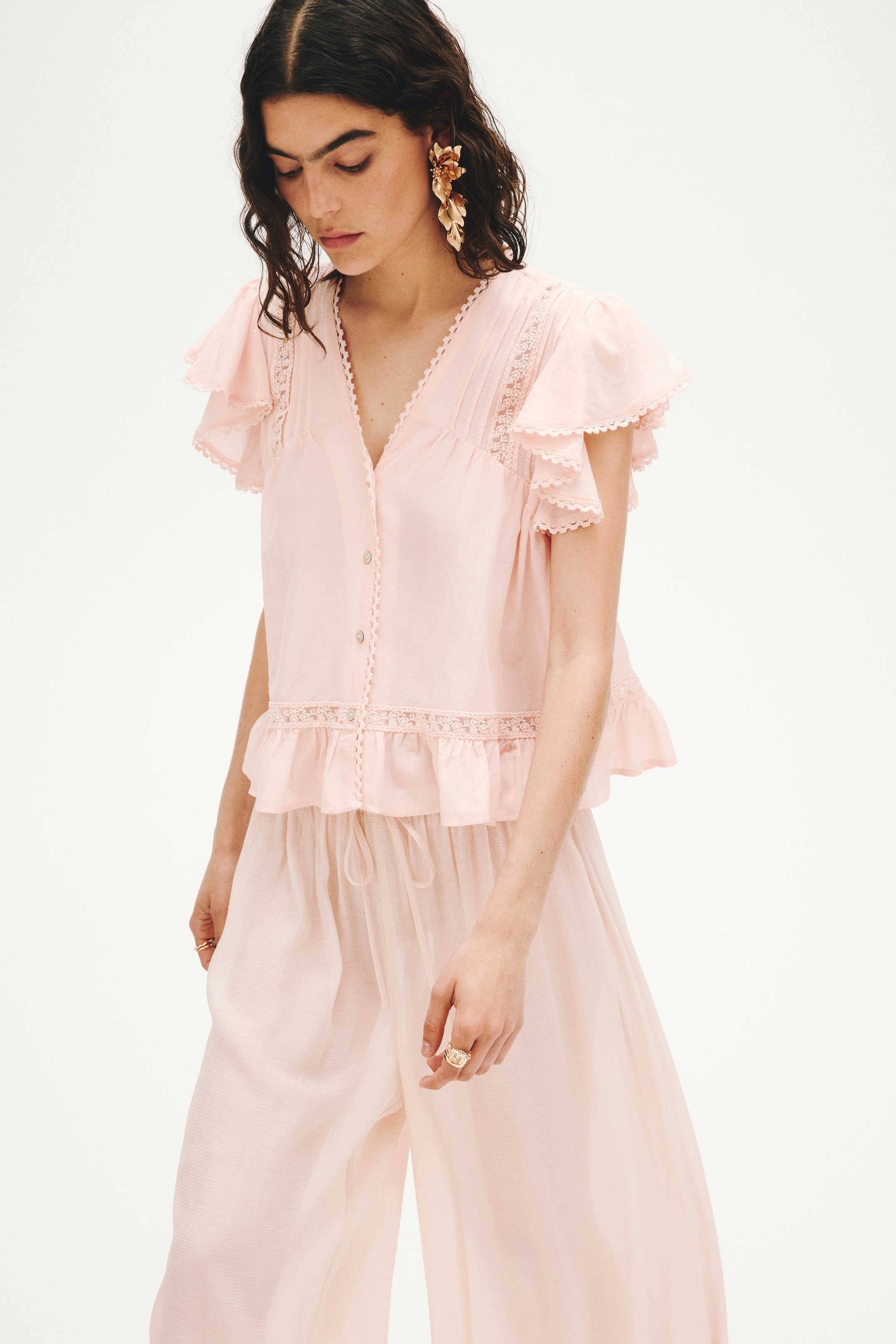 Ver imagen más grande: La modelo lleva una blusa rosa claro en un tejido ligero, con un escote en V profundo, botones en la parte delantera, mangas cortas de mariposa, inserciones de encaje, pliegues y un dobladillo con volantes. Se combina con un pantalón rosa claro a juego.